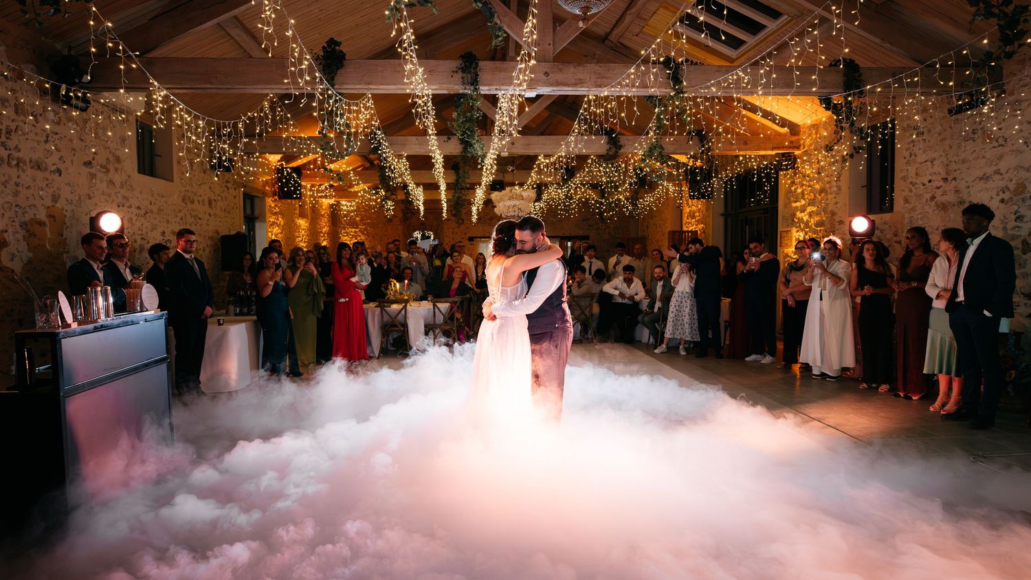 Bride and groom kiss during their first dance in a cloud of dancing fog under twinkling string lights in a stone wedding barn.