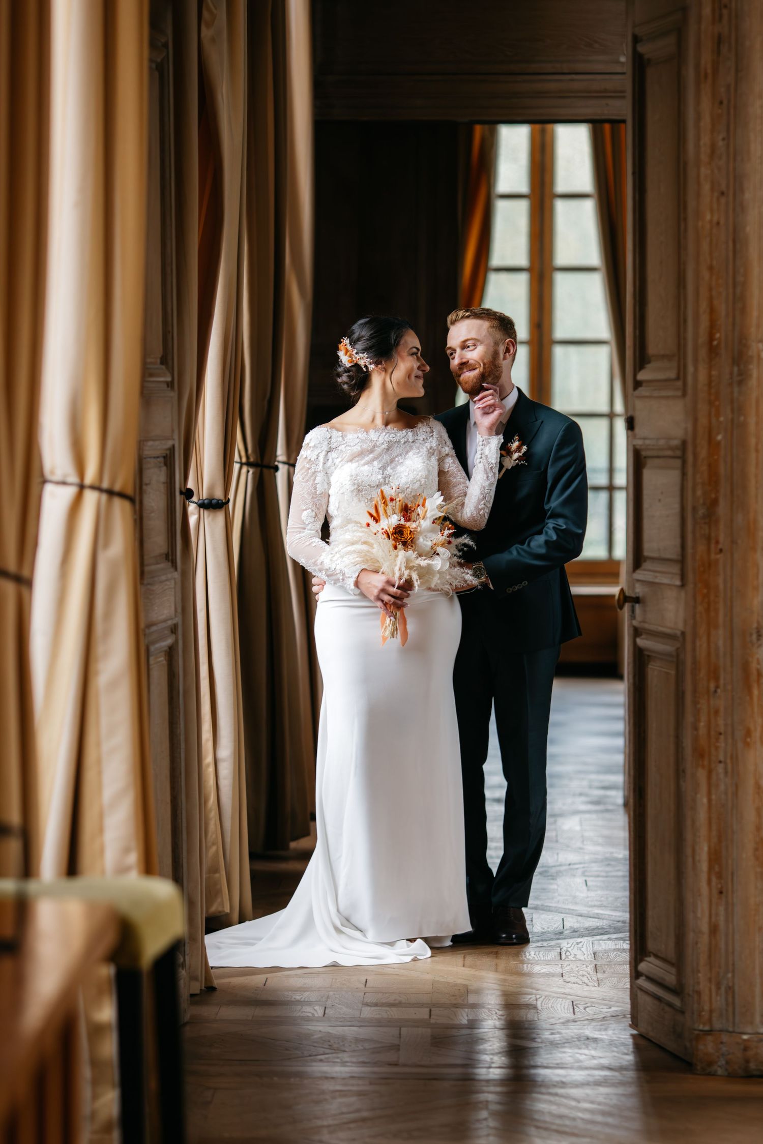 Bride in a lace-sleeved gown and groom in a navy suit embrace in an elegant doorway with a bouquet of dried flowers.