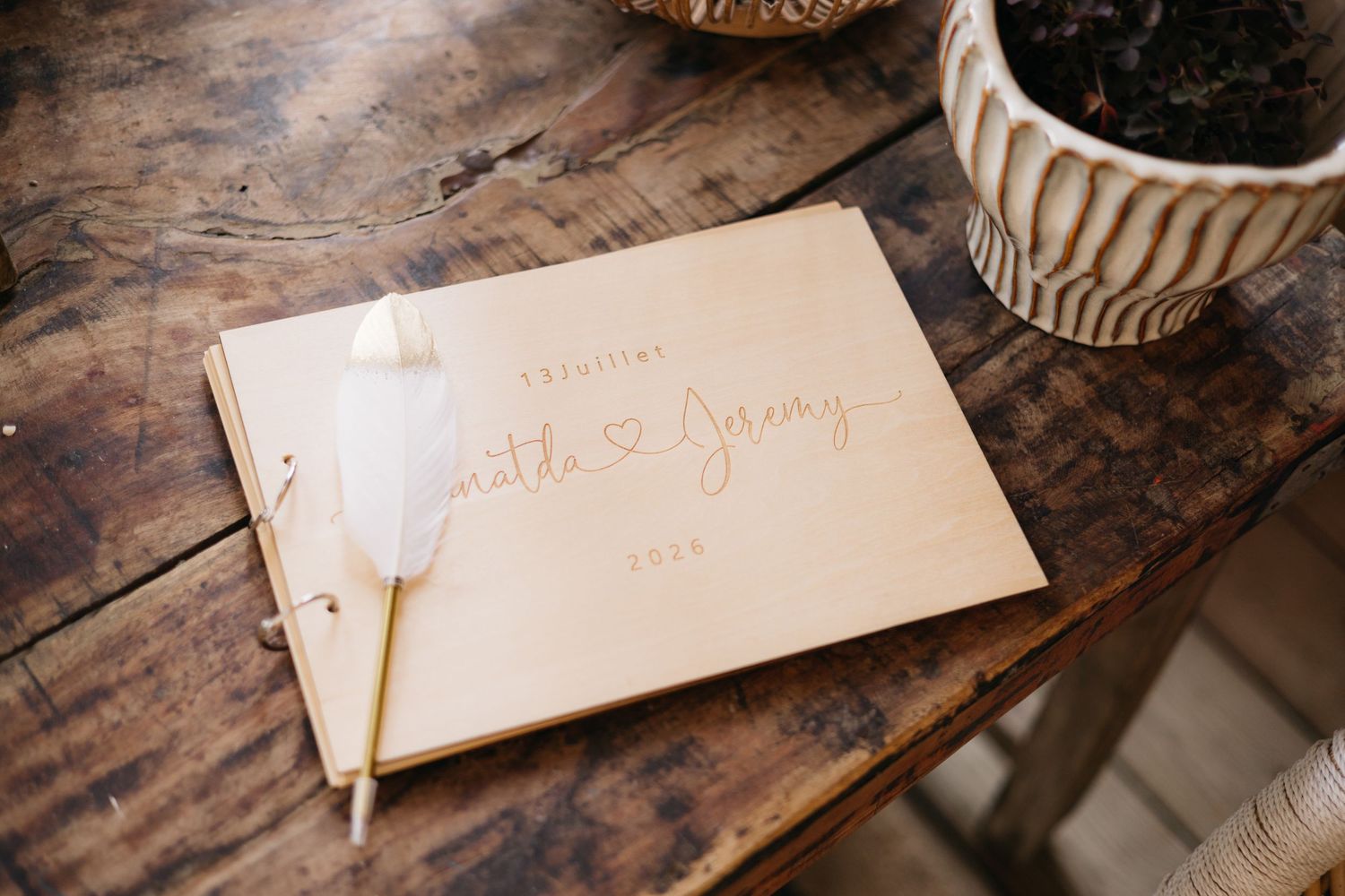 Livret de invités de mariage en bois avec les noms des mariés gravés et un stylo à plumes blanches reposant sur une table en bois rustique.