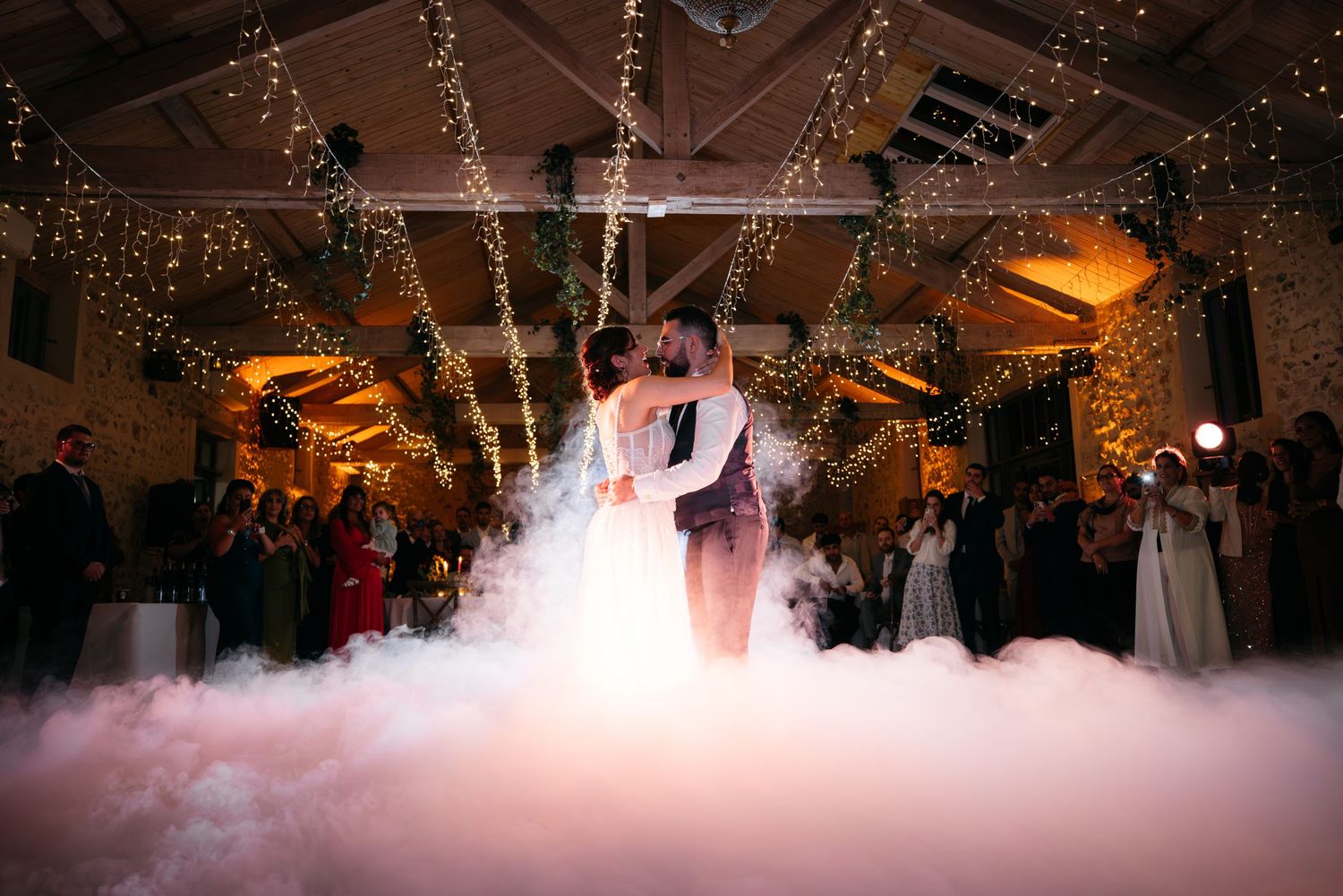 Couple kisses in a glowing haze of dance floor fog while wedding guests look on beneath a canopy of warm fairy lights.