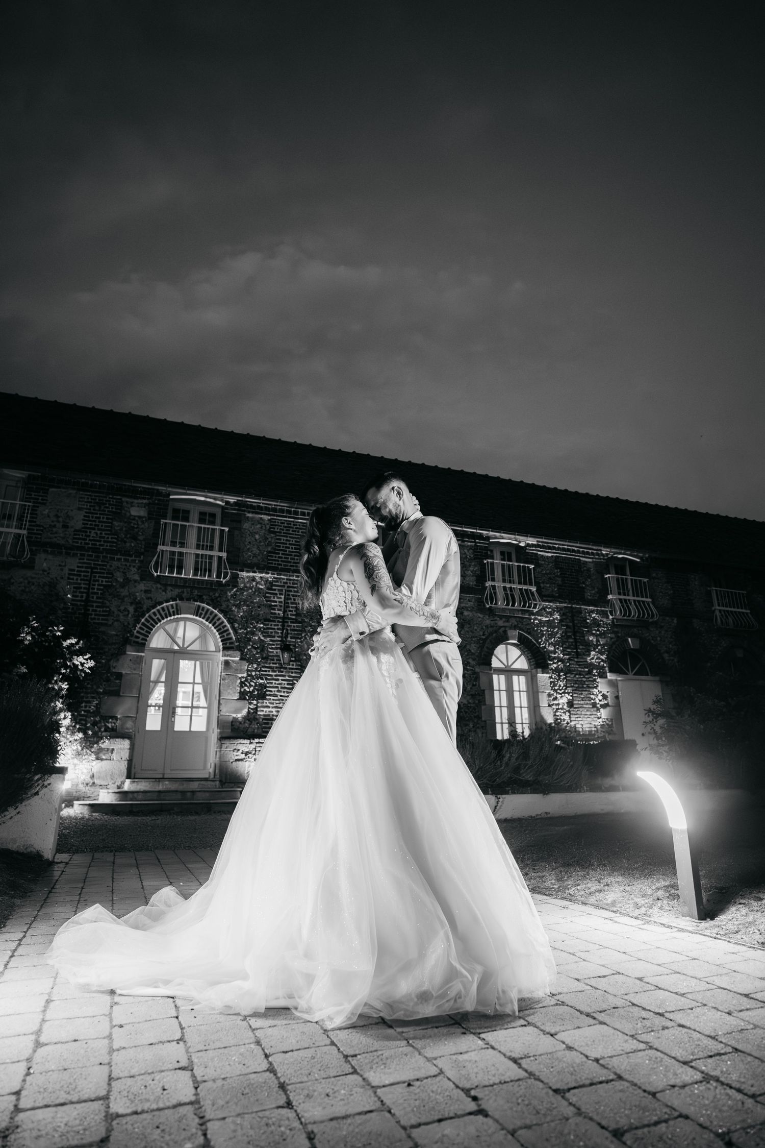 Photo en noir et blanc d'une mariée dans une robe en tulle complète embrassant son mari sur une cour en pierre la nuit