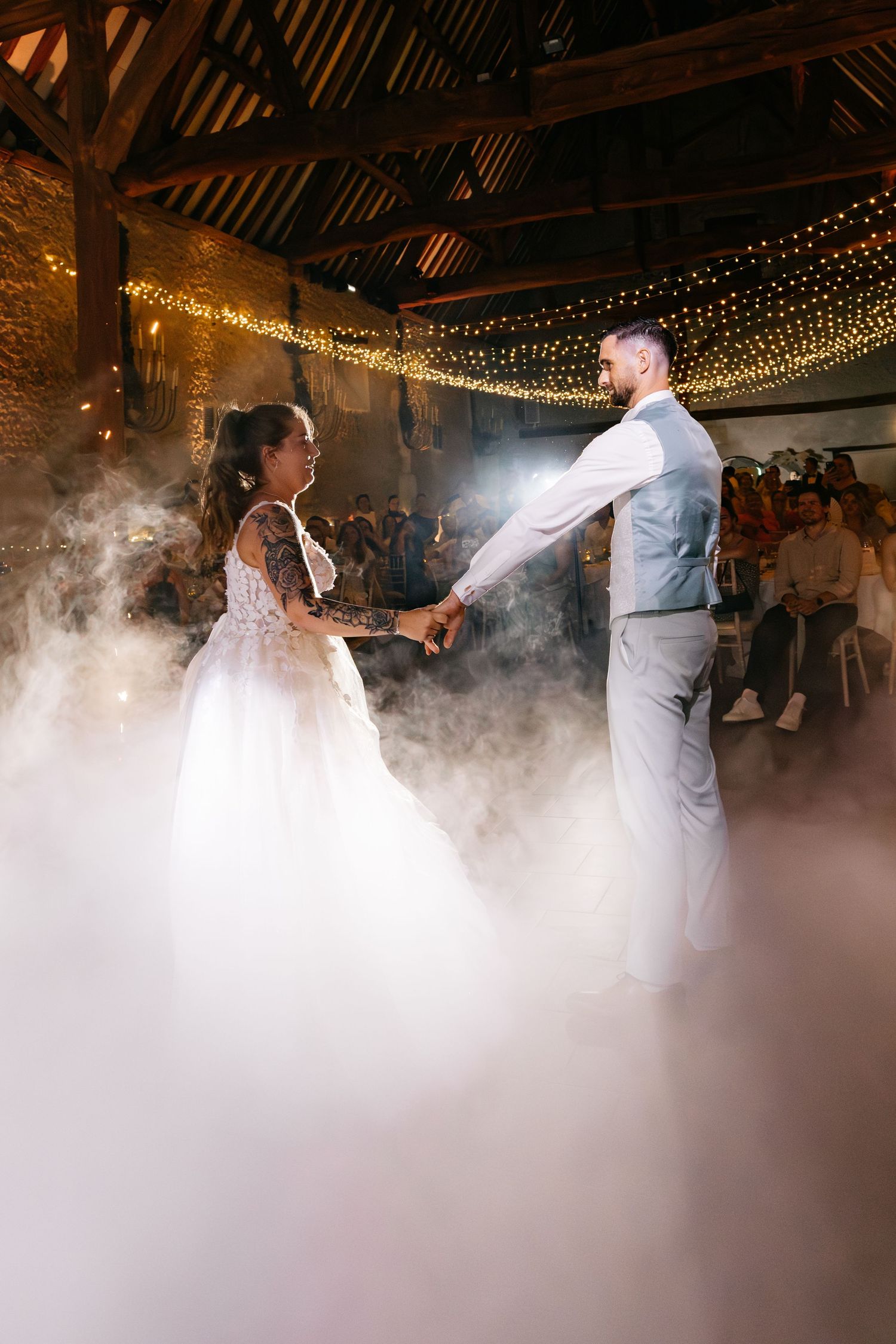 La mariée avec un bras tatoué et le marié dans une veste se tiennent la main pour leur premier danse dans un brouillard bas dans une grange