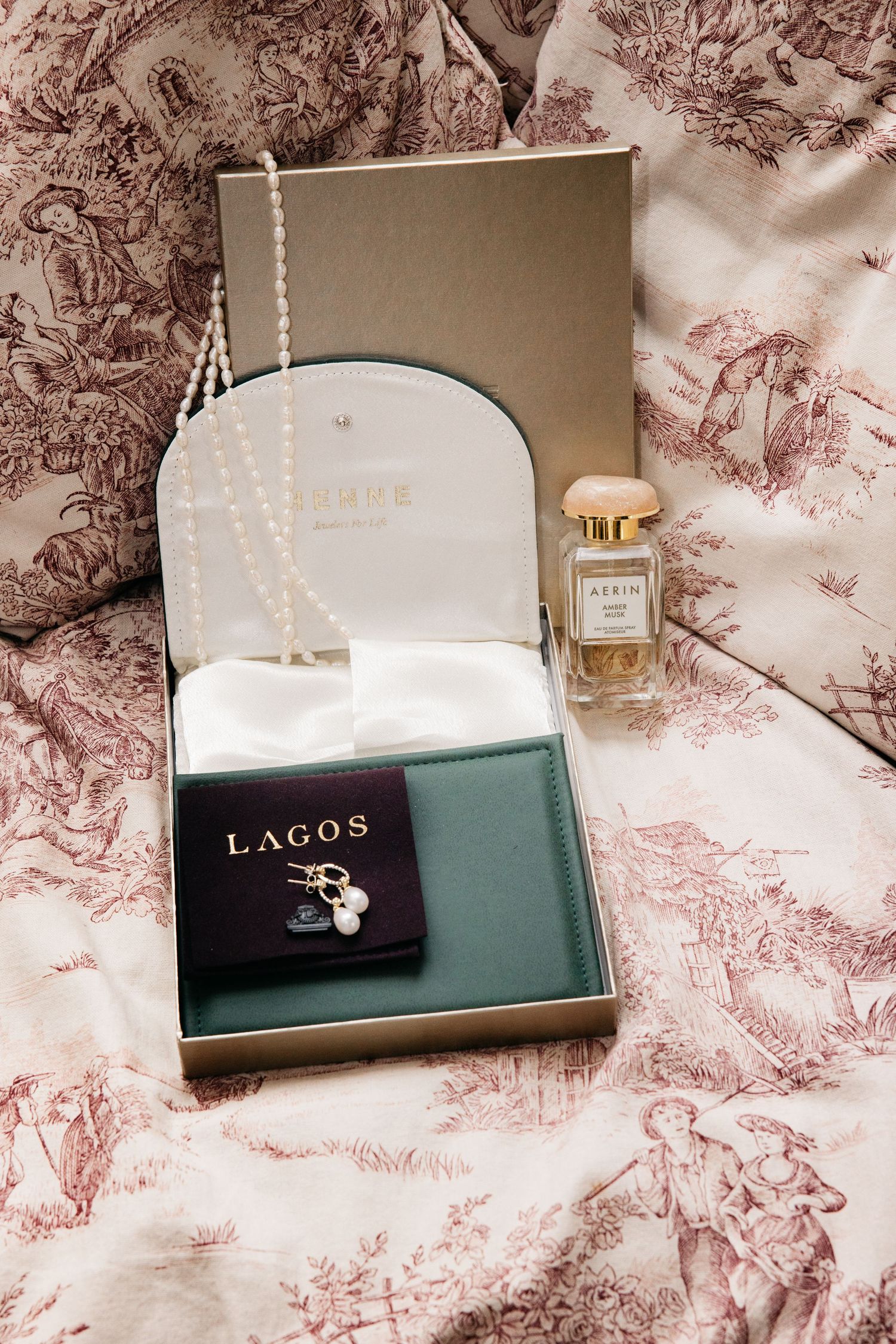Open jewelry box with a pearl necklace, Lagos earrings pouch and a perfume bottle resting on pink toile bedding.