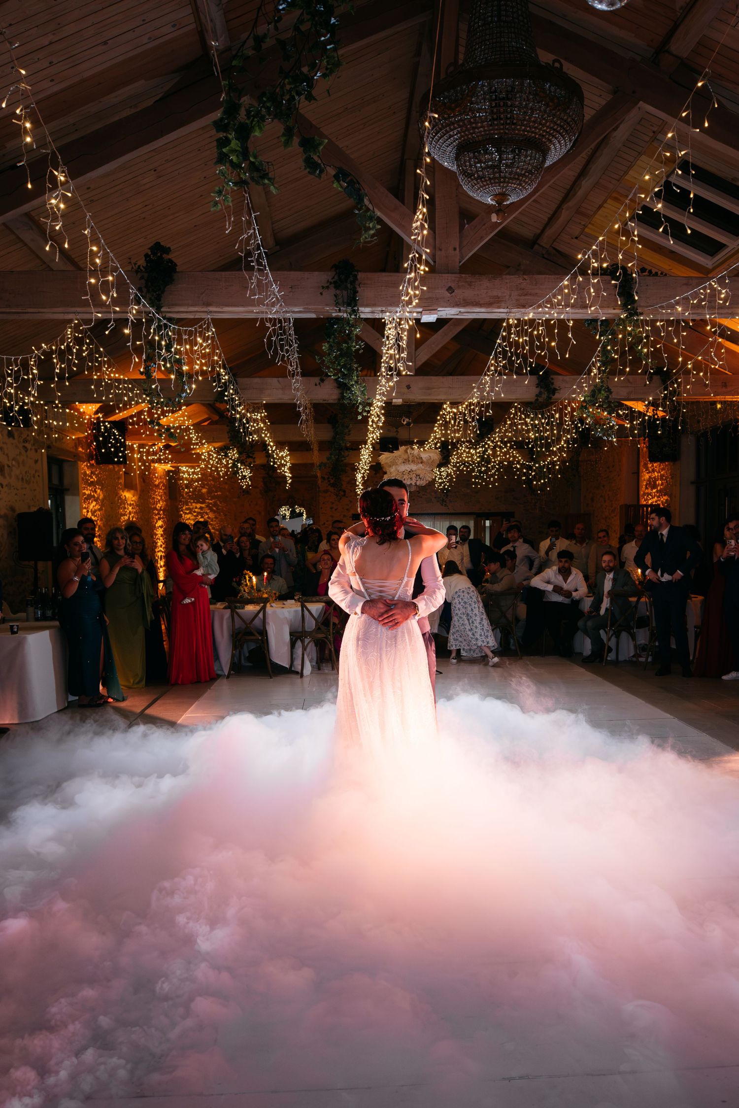 Bride in a flowing white gown embraces her partner on a fog-covered dance floor beneath wooden beams, fairy lights, and a chandelier.