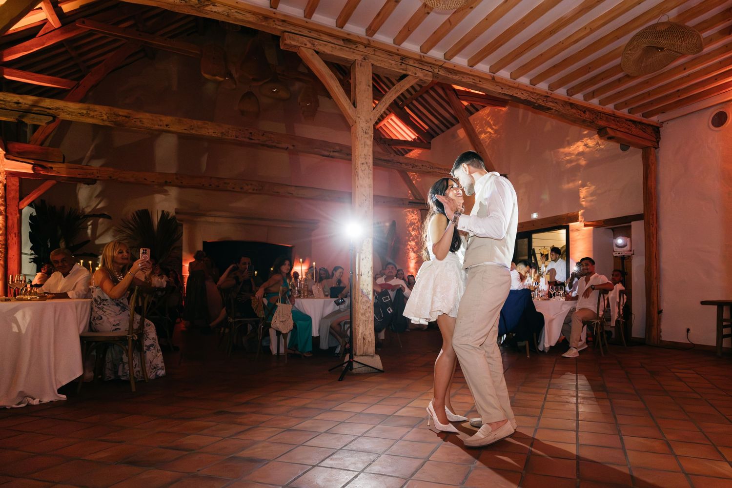 Newlyweds dance together in a warmly lit rustic barn venue with exposed wooden beams while seated wedding guests look on.