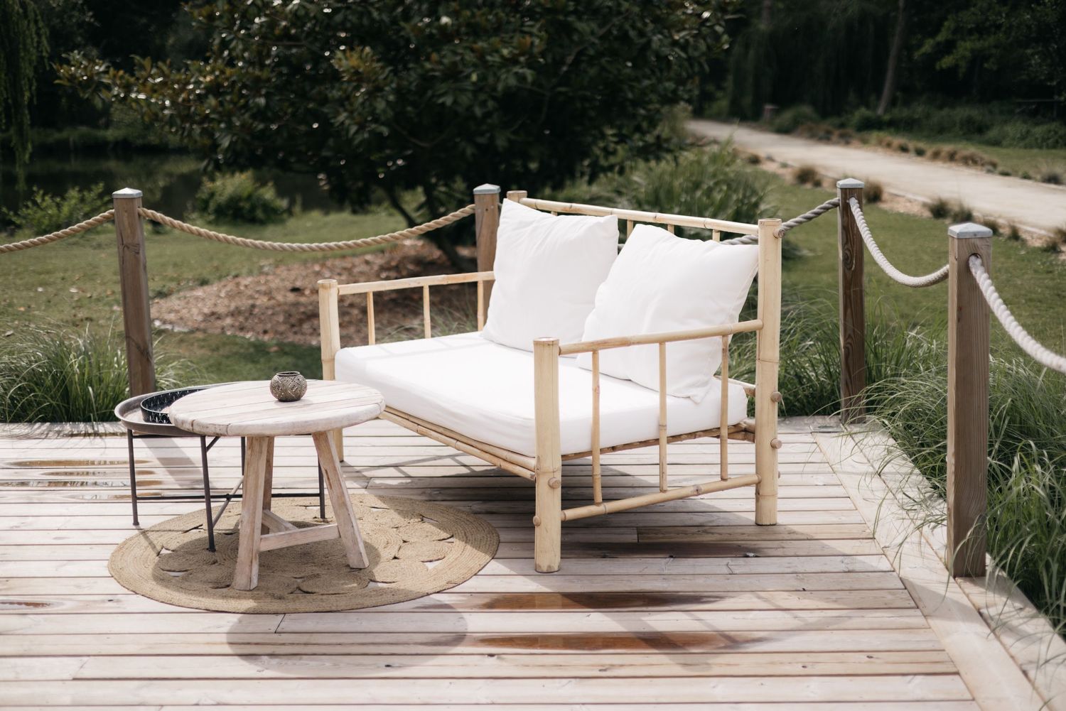 White cushioned wooden daybed with a round side table on a wooden deck surrounded by greenery at an outdoor wedding venue.