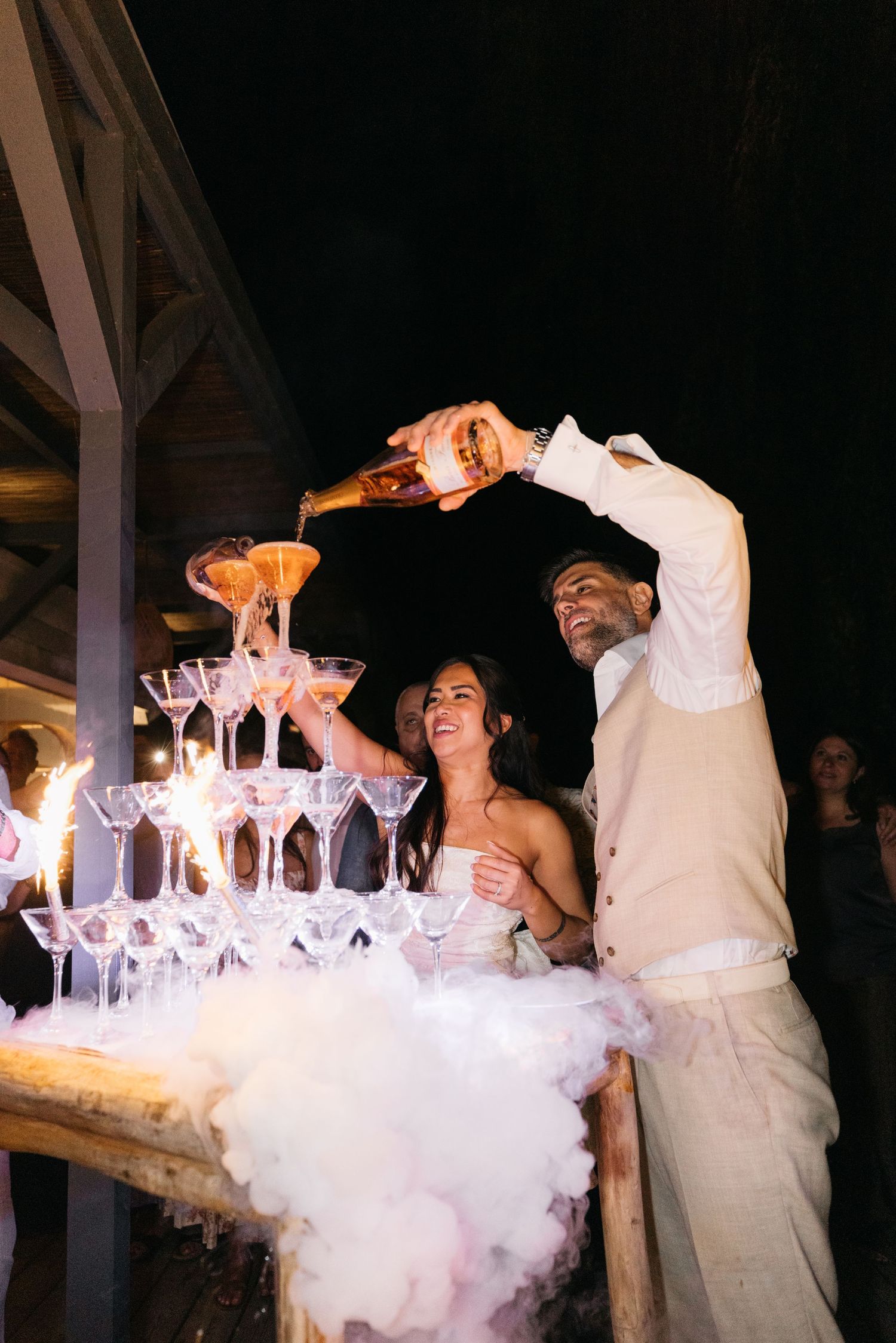 La mariée et le marié versent du champagne rose sur une tour de verres à coupe avec des effets fumants et des paillettes