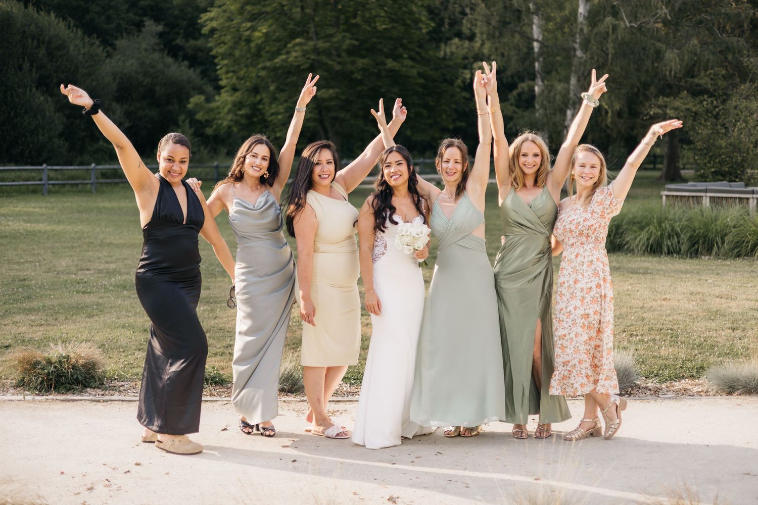 Bride poses with six bridesmaids in pastel dresses, arms raised in celebration, on a sunny lawn at an outdoor wedding venue.