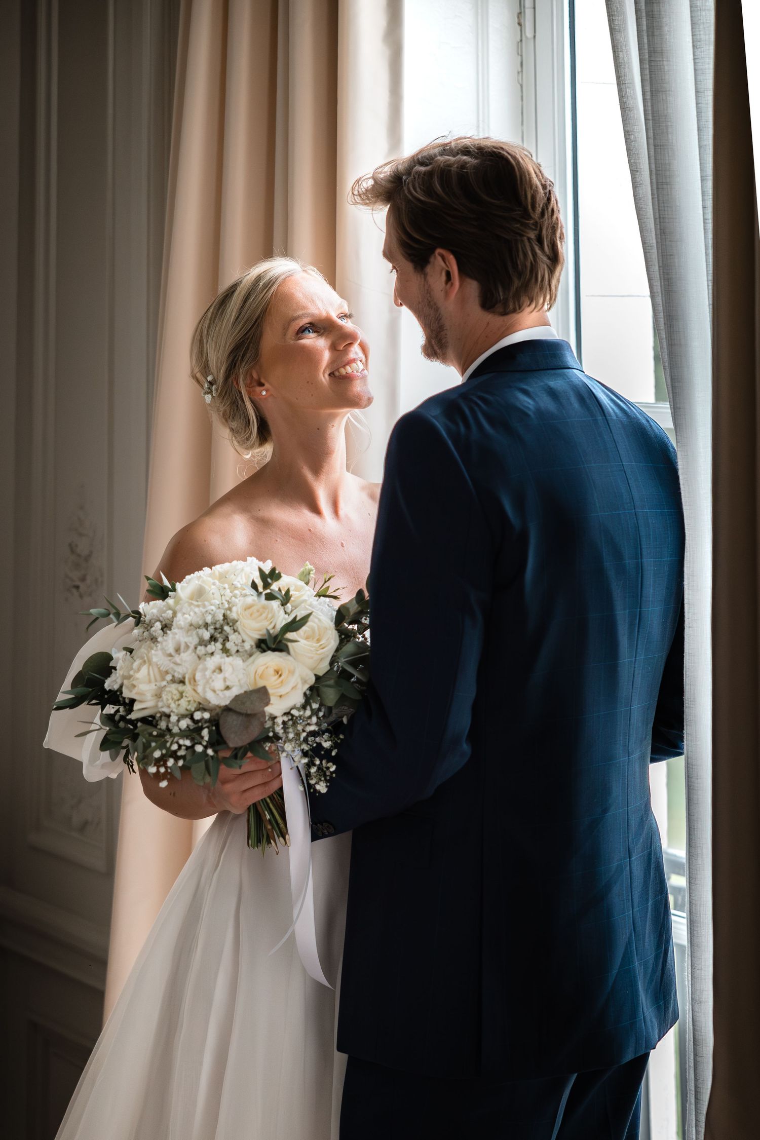 Smiling bride holding a white rose bouquet gazes at her groom in a navy suit beside a sunlit window.
