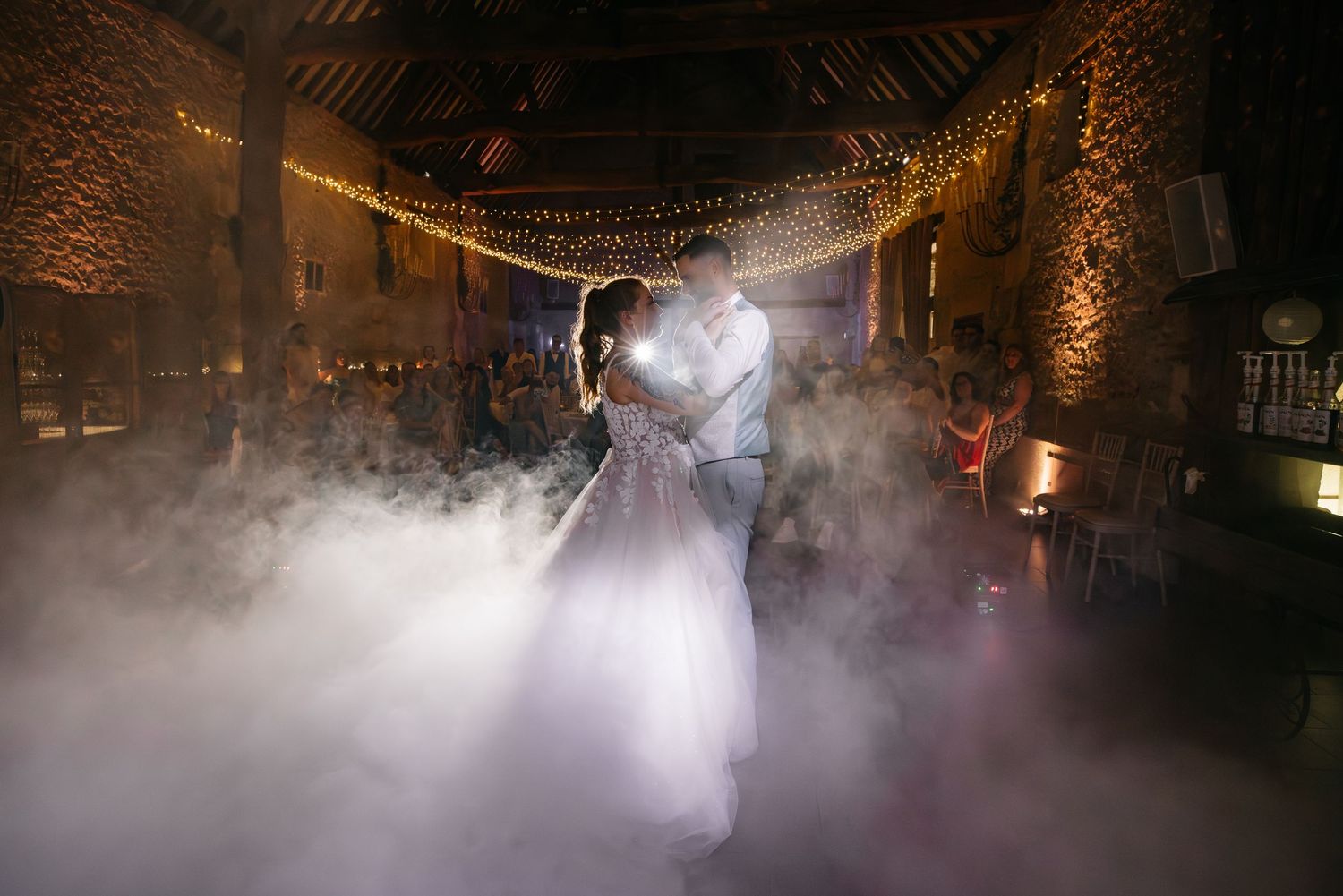 La mariée et le marié dansent près l'un de l'autre dans une brume tourbillonnante sous des poutres en bois rustiques et des guirlandes