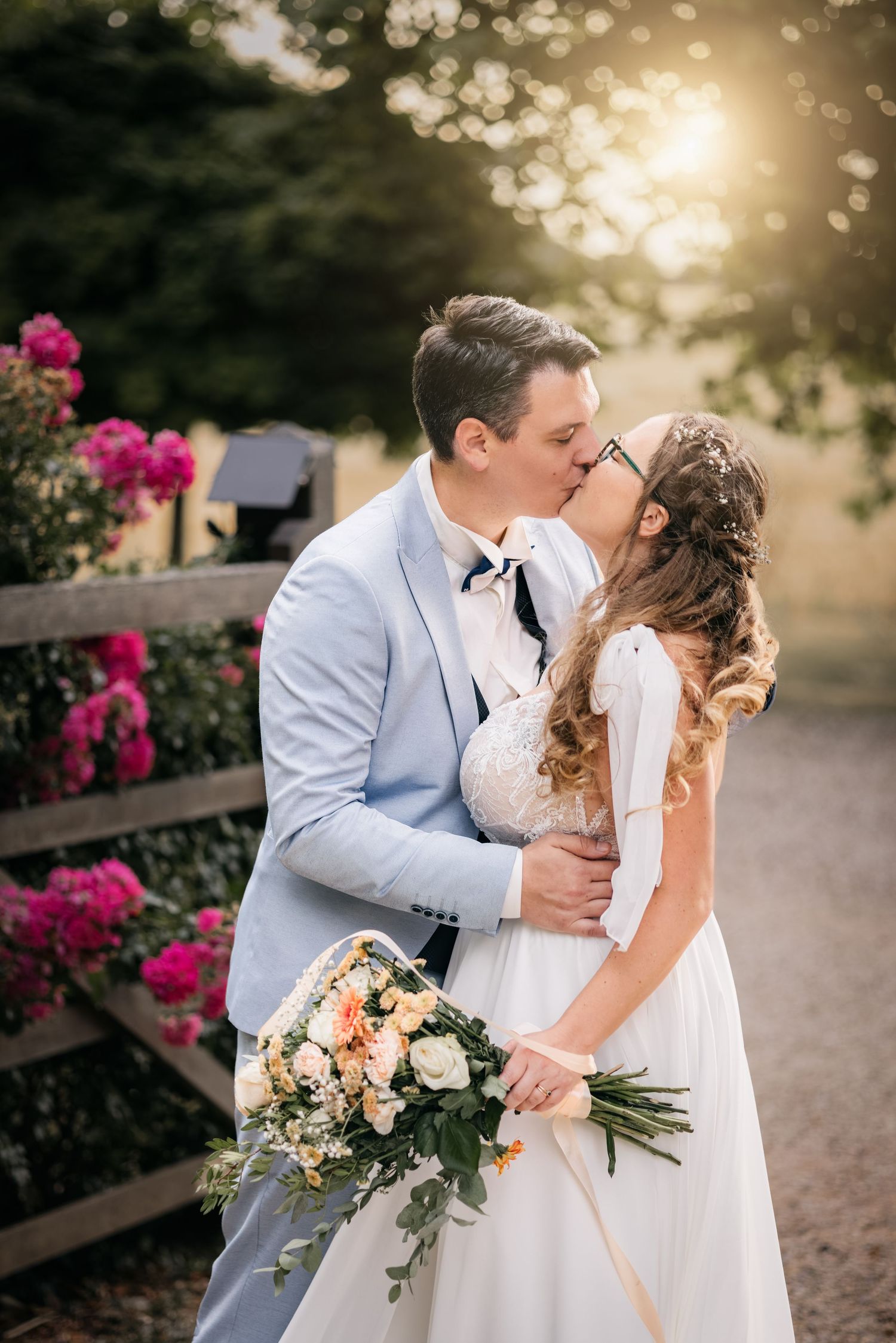 Bride and groom share a kiss in golden sunlight beside pink roses, the bride holding a bouquet of cream and peach flowers.