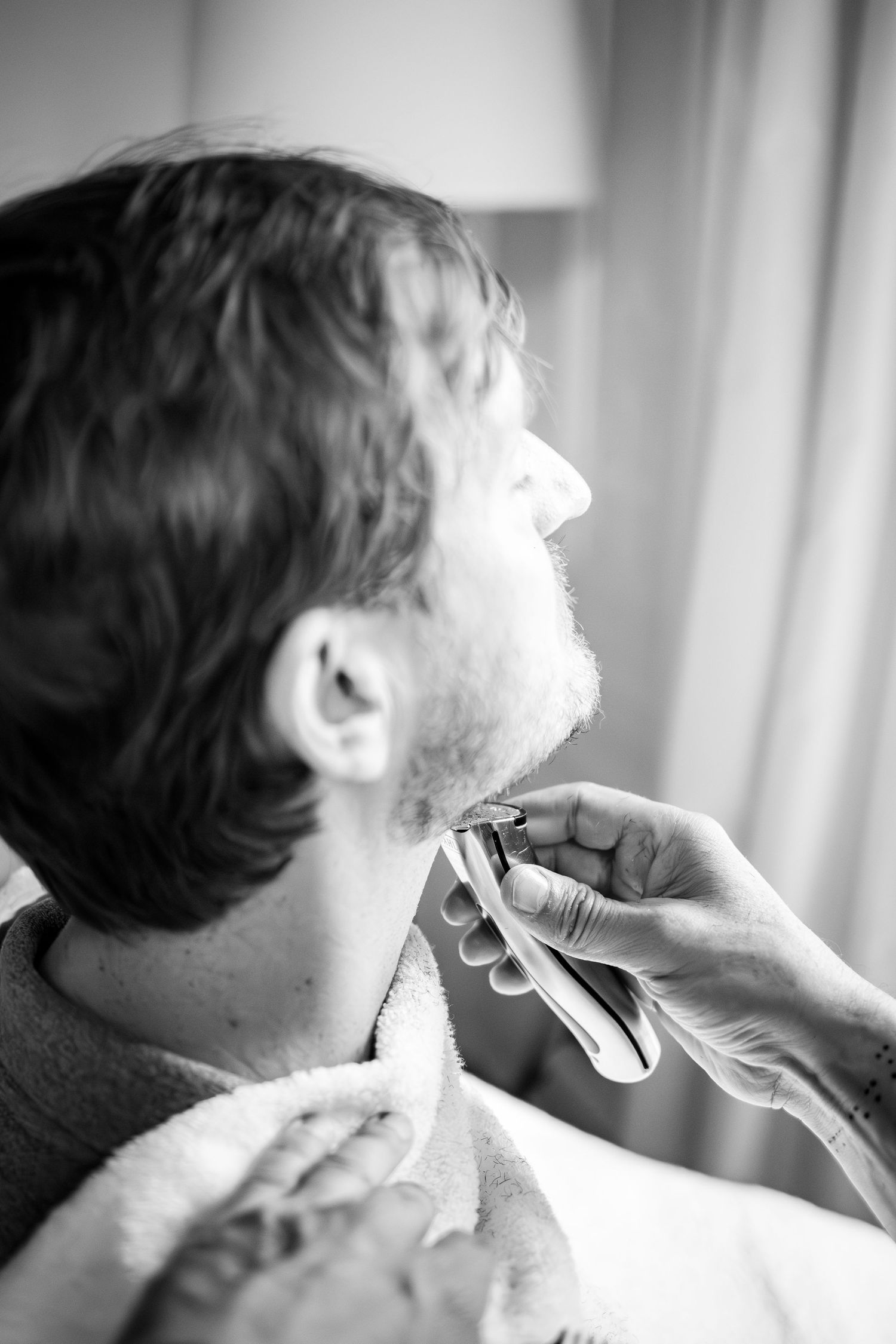 Black and white photo of a groom getting ready as a barber carefully shaves his beard with a razor.