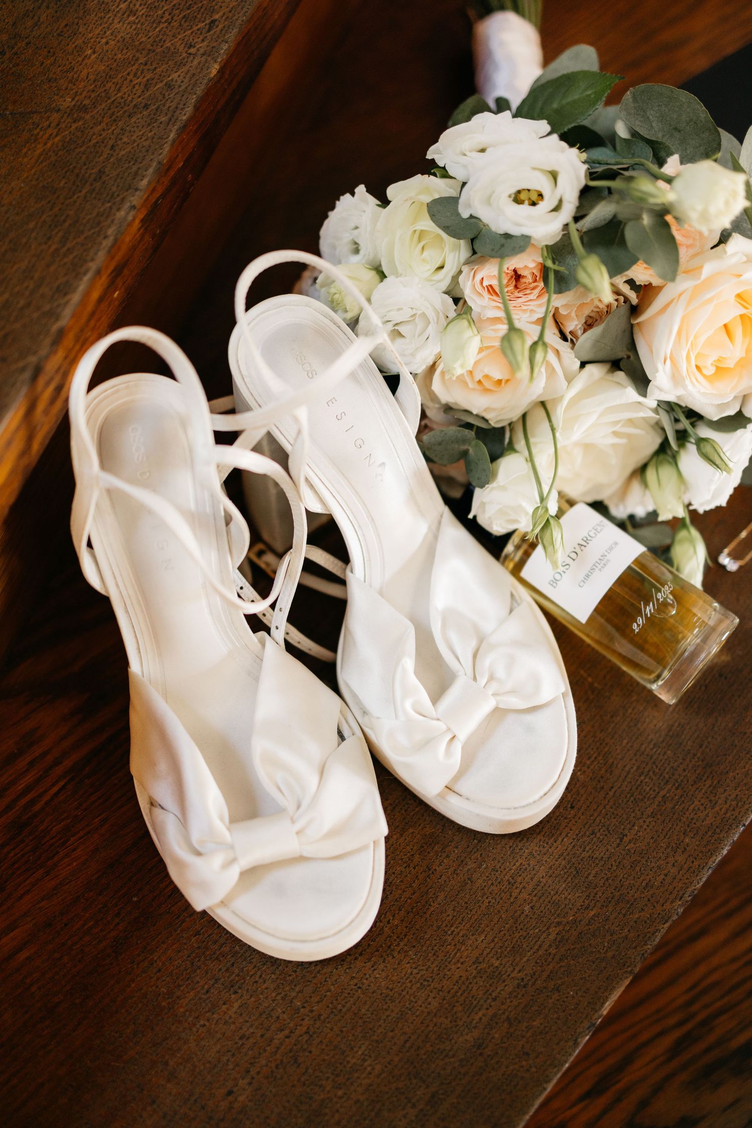 White strappy bow sandals beside a bridal bouquet of cream and blush roses and a perfume bottle on a dark wooden surface.