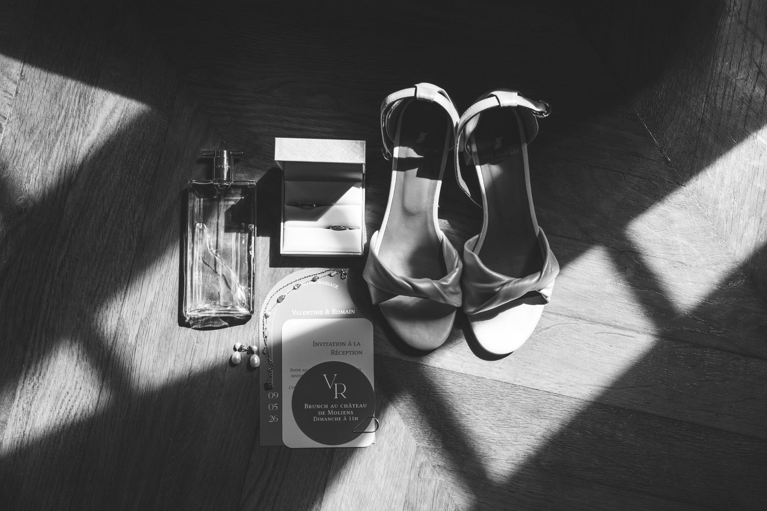 Black and white flat lay of bridal sandals, perfume bottle, and wedding details on a wooden floor with window shadows.