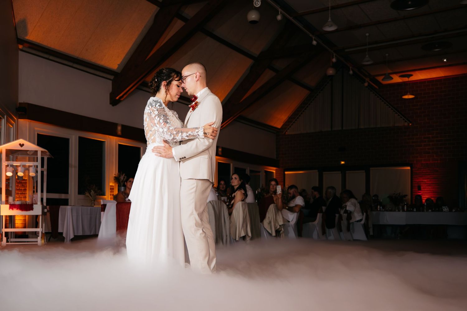 Newlyweds share their first dance wrapped in low dry-ice fog in a rustic wooden-beamed reception hall while guests watch from the tables.