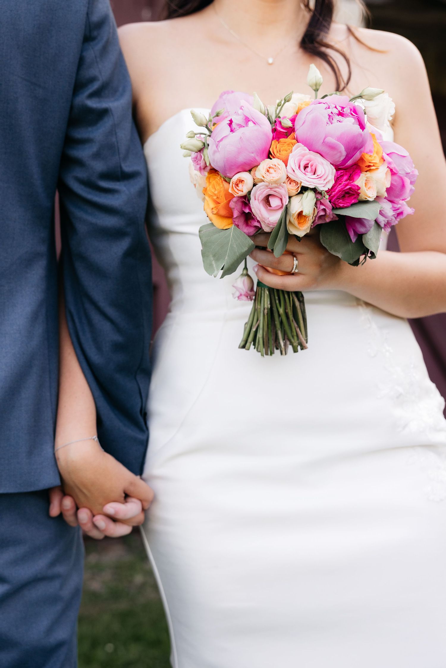 Bride in a white strapless gown holds a pink and orange peony bouquet while holding hands with the groom in a blue suit.