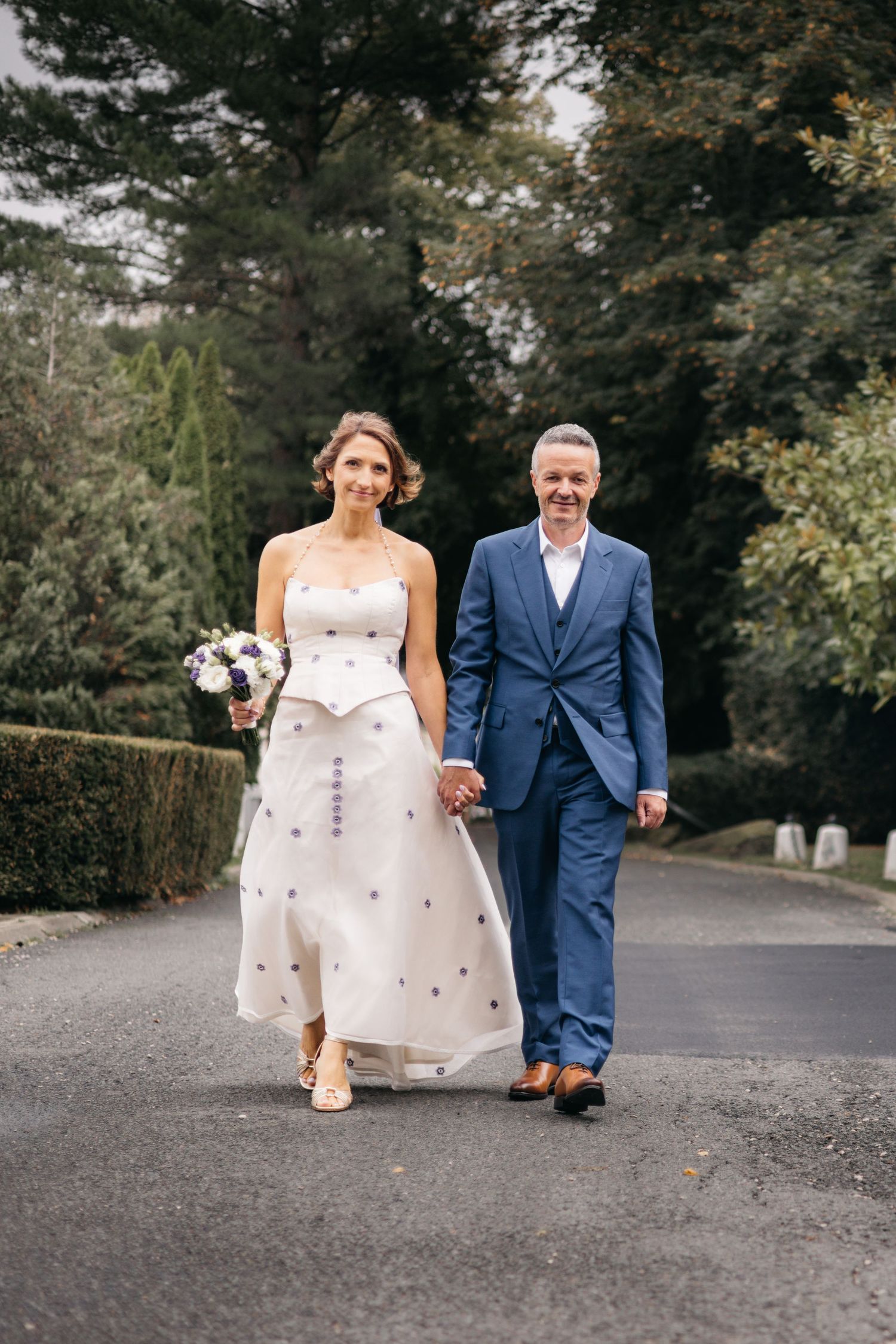 Bride in a white dress with a bouquet and groom in a blue suit walk hand in hand along a tree-lined garden path.