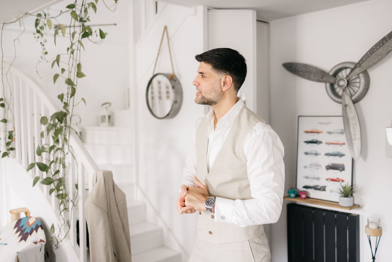 Groom in a cream vest and white shirt stands by a staircase with trailing plants, looking thoughtfully into the distance.