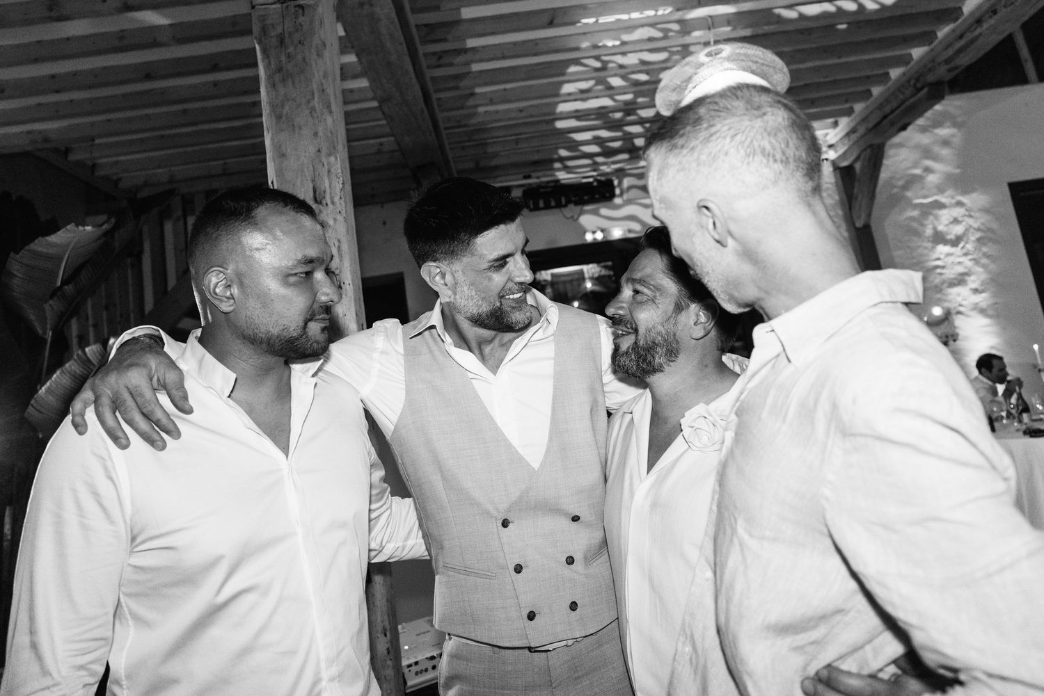 Photo en noir et blanc du marié qui rit avec ses compagnons de cérémonie dans une étreinte candide lors d'une célébration de mariage.
