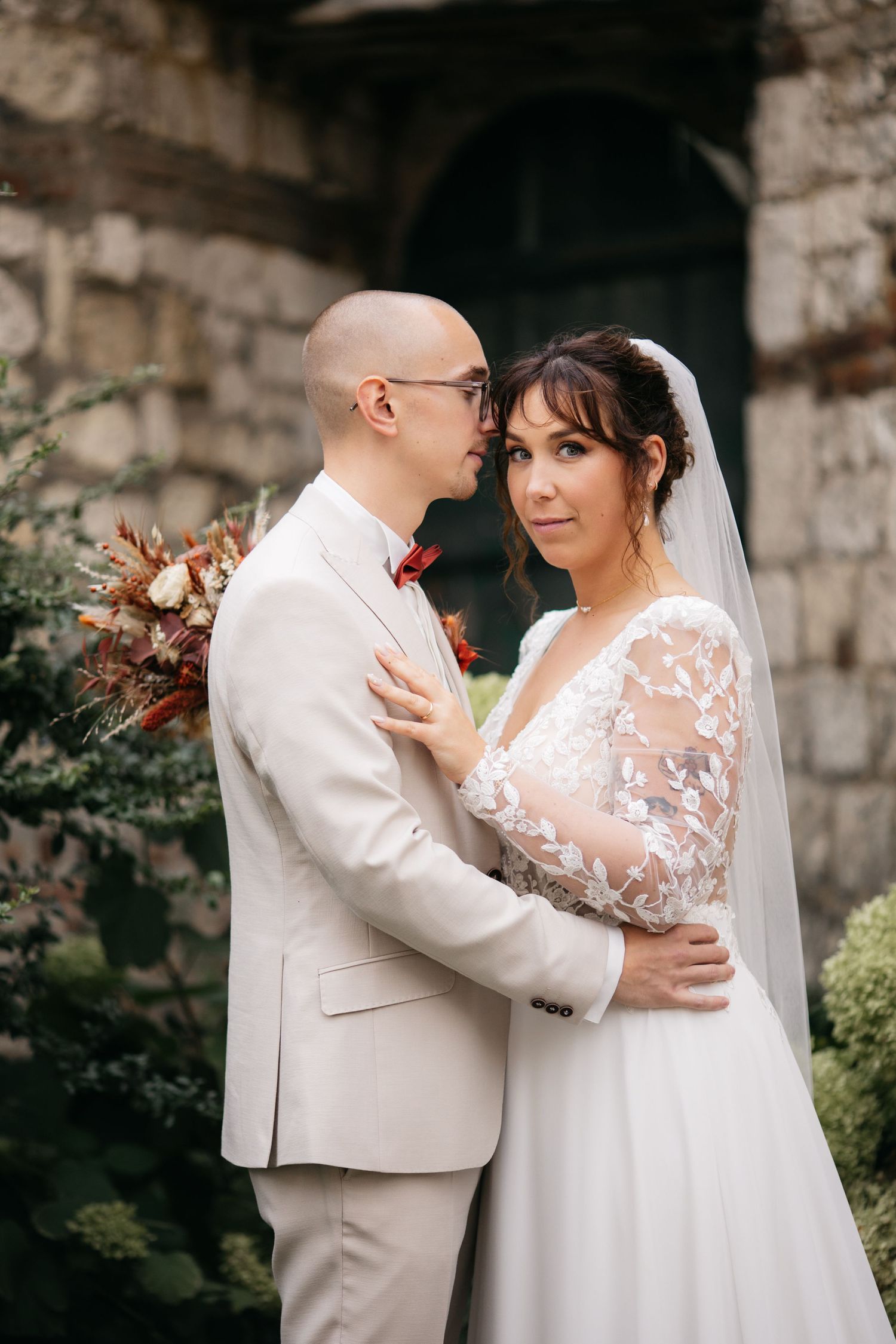Bald groom with glasses and a beige suit embraces a bride in a lace-sleeved wedding gown and veil beside a stone wall.