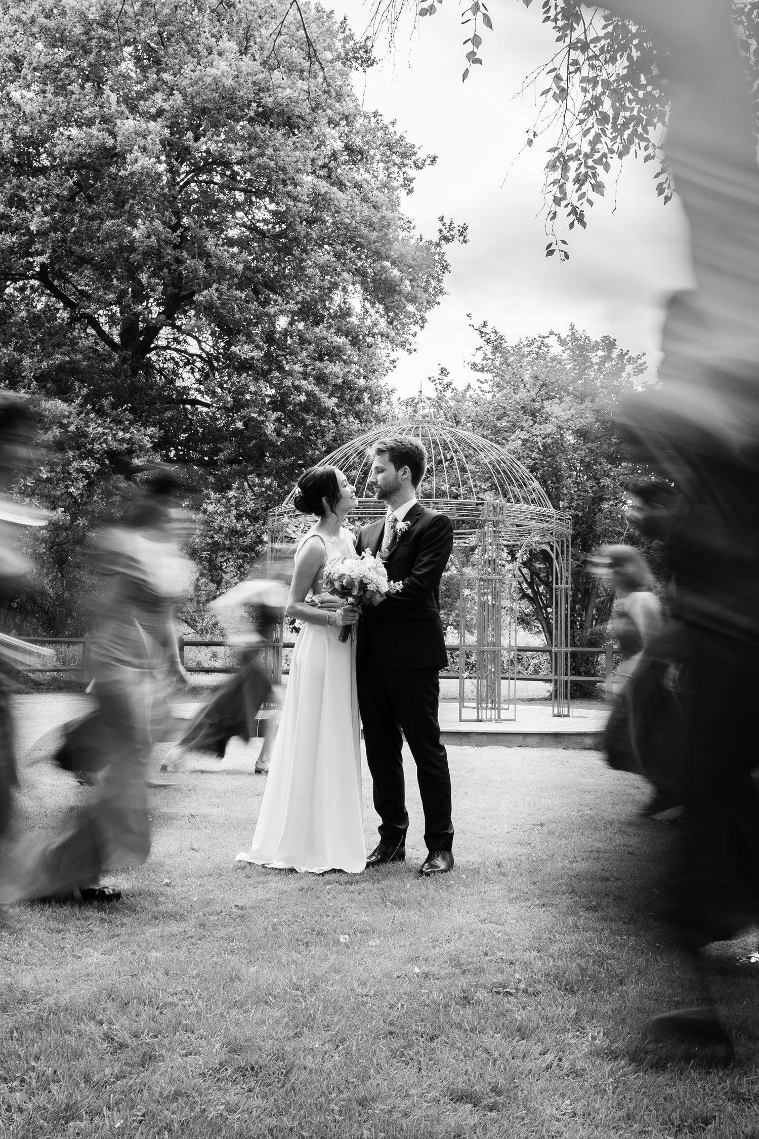 Black and white wedding photo of a bride and groom embracing near a garden gazebo while blurred guests move around them.