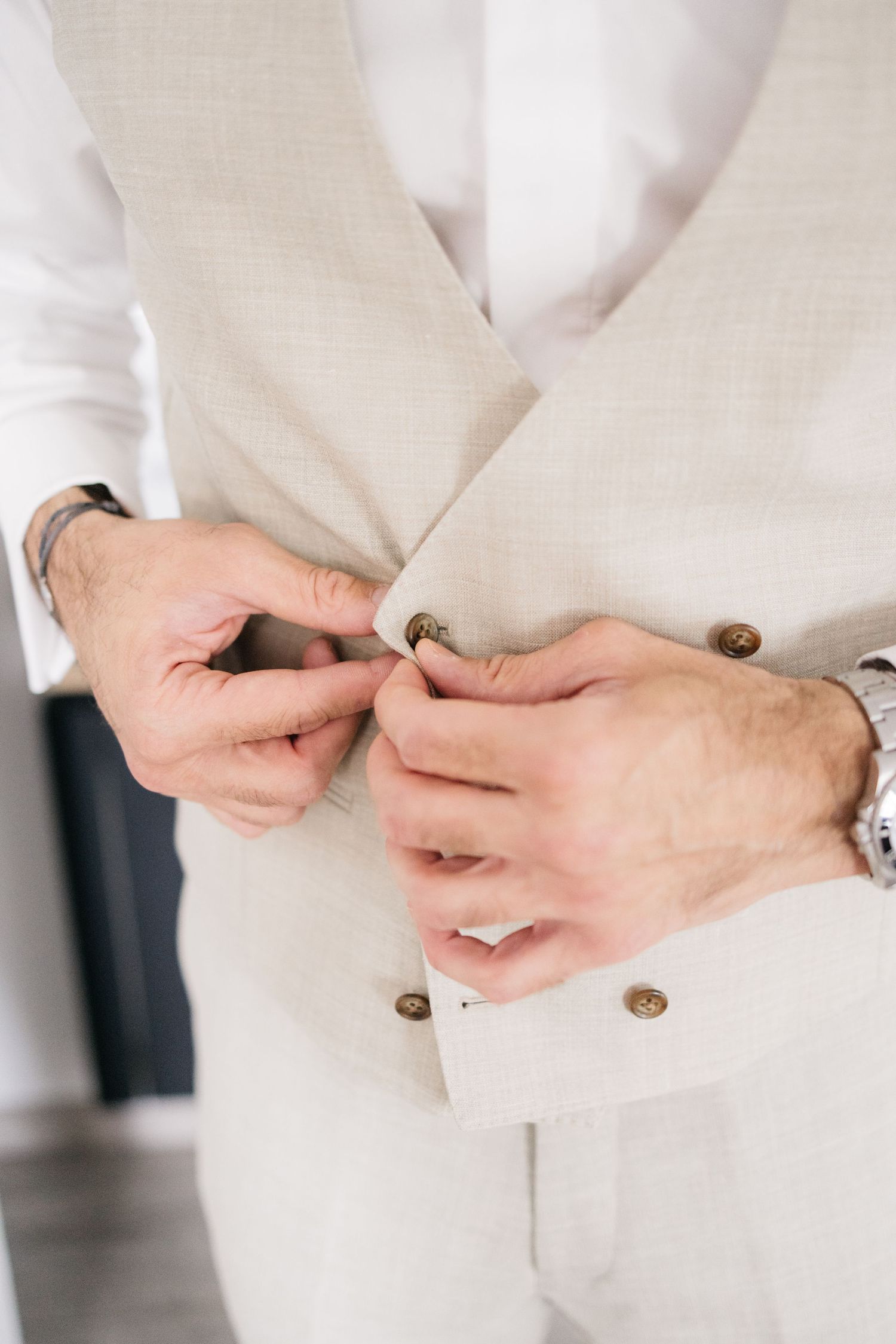 Groom in a cream double-breasted linen vest buttoning it over a white dress shirt while getting ready for the wedding.