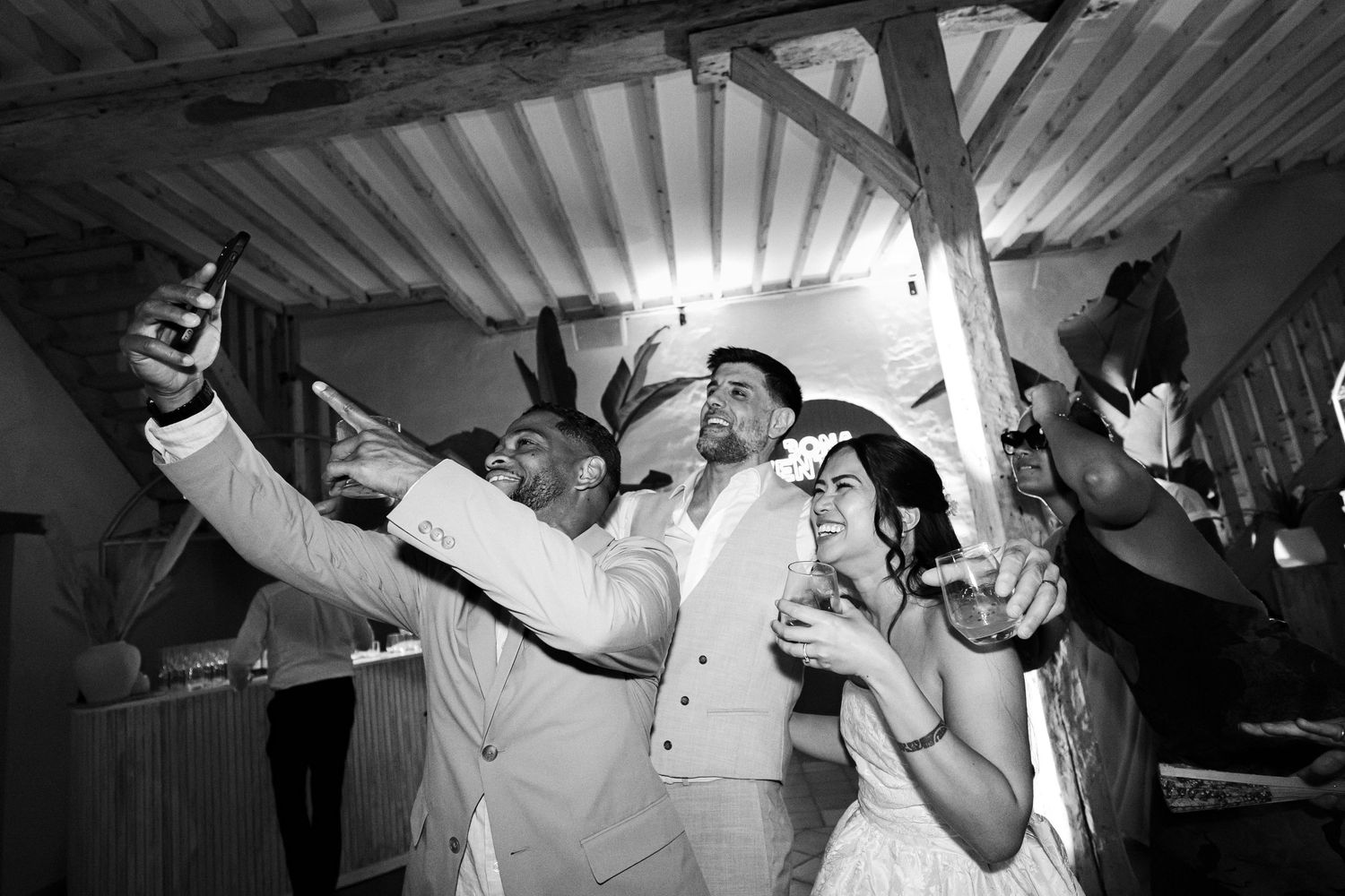 Selfie en noir et blanc de la mariée, du marié et des amis qui rient ensemble lors d'une réception de mariage animée.