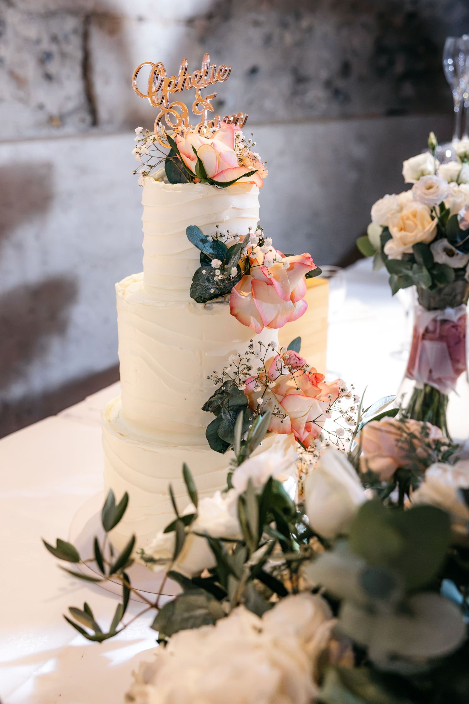 Elegant white two-tier wedding cake decorated with pink roses, greenery, and a gold cake topper, surrounded by white floral centerpieces.