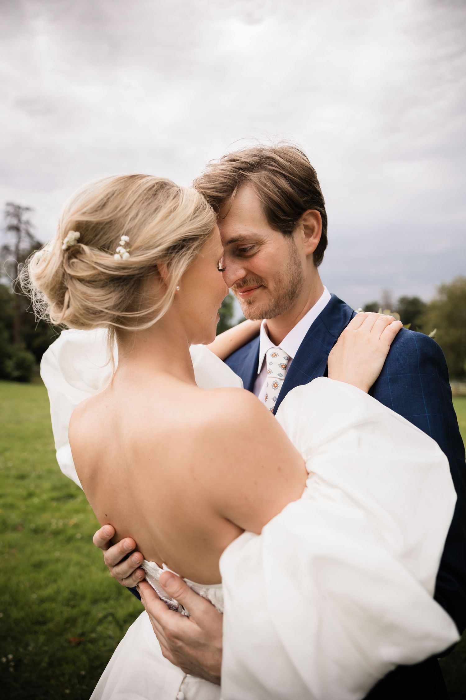 Bride and groom share a tender forehead-to-forehead moment outdoors on a green lawn under a cloudy sky.