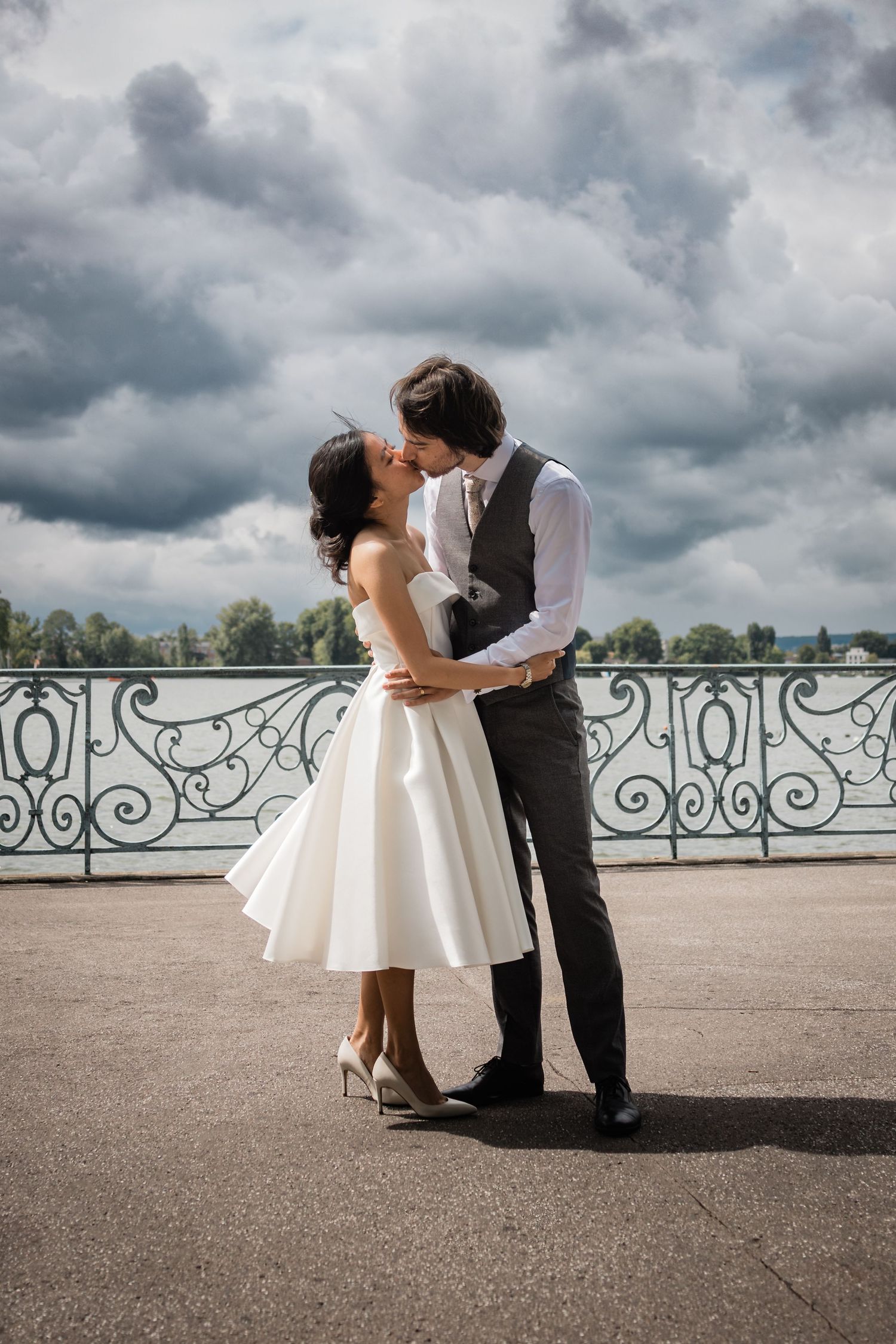 Bride in a short white tea-length dress and groom in a gray vest kiss on a waterfront terrace beneath a dramatic cloudy sky.
