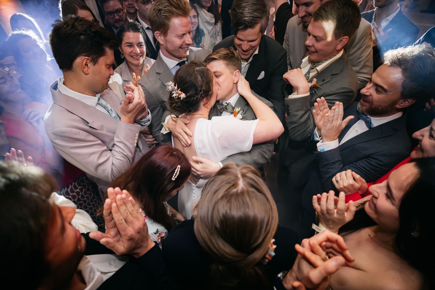 Wedding guests crowd around the newlyweds, clapping and cheering as the couple shares a joyful kiss on the dance floor.
