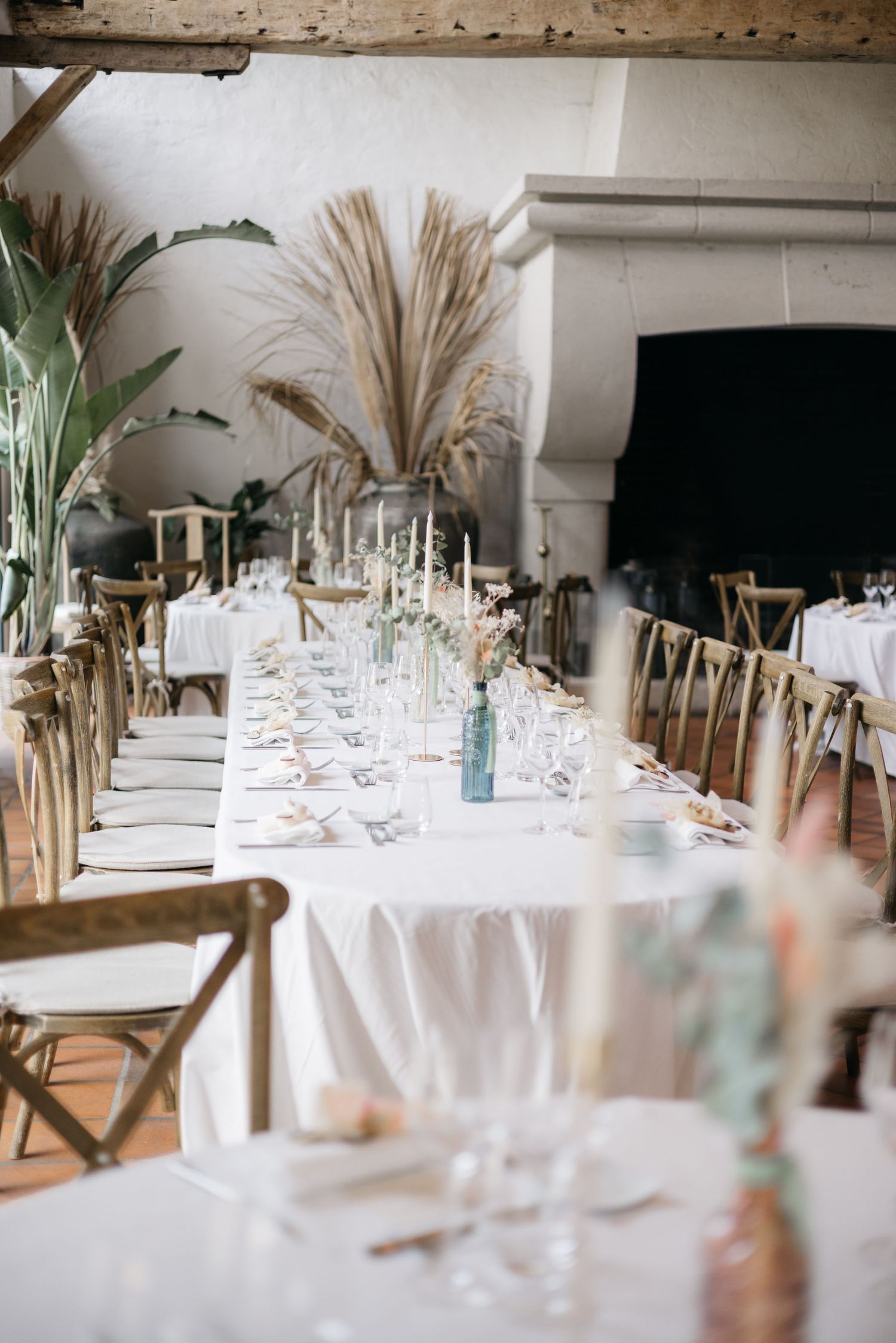 Long rustic wedding banquet table with white linens, glassware, candles and dried pampas grass decor beside a stone fireplace.