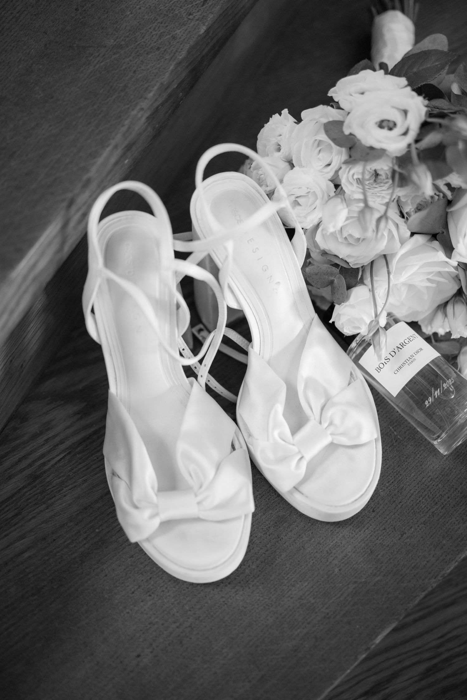 Black and white photo of white bow-detail wedding sandals beside a bouquet of roses and a perfume bottle on a wooden floor.