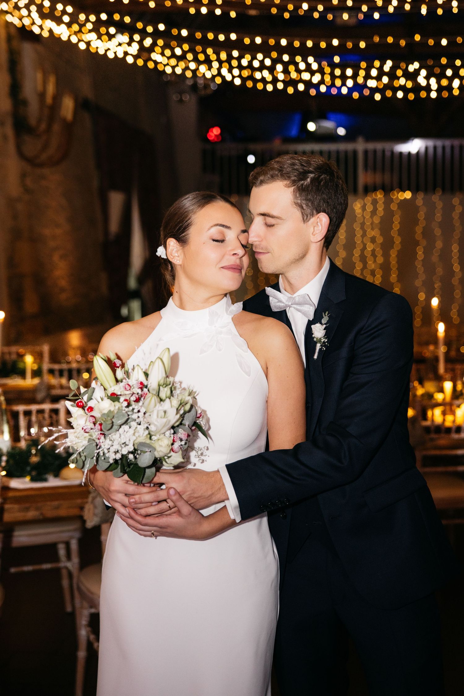 Bride in a halter gown with a white bouquet is embraced by her groom in a black tuxedo under warm string lights at an evening reception.