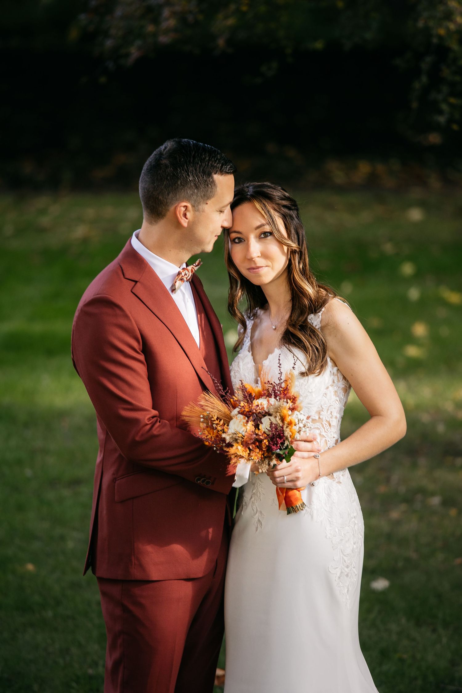 Groom in a rust-red suit and bow tie leans toward his smiling bride in a lace dress holding an autumn bouquet outdoors.