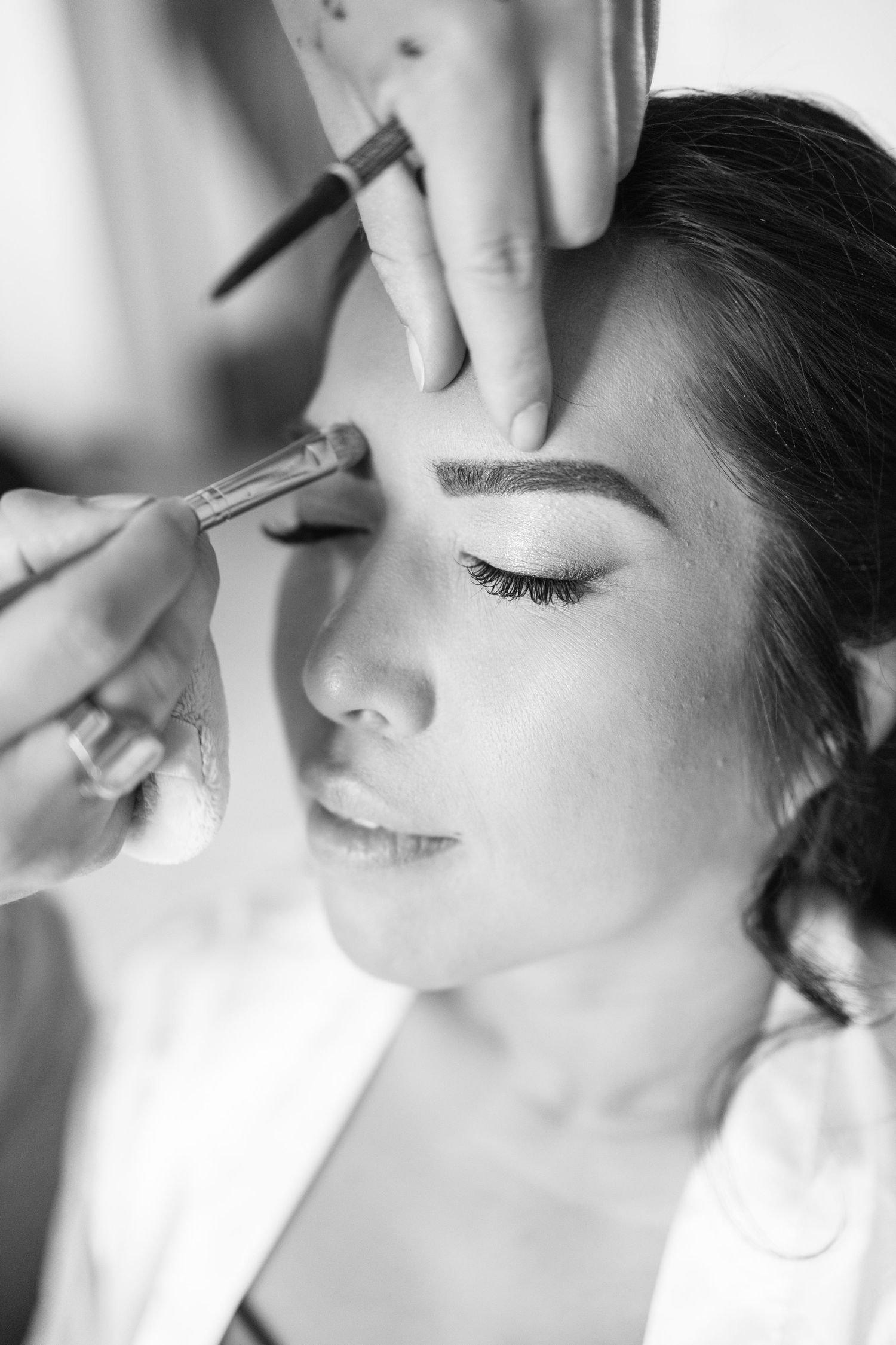Black and white close-up of a bride with closed eyes as a makeup artist shapes her eyebrows with a brush.