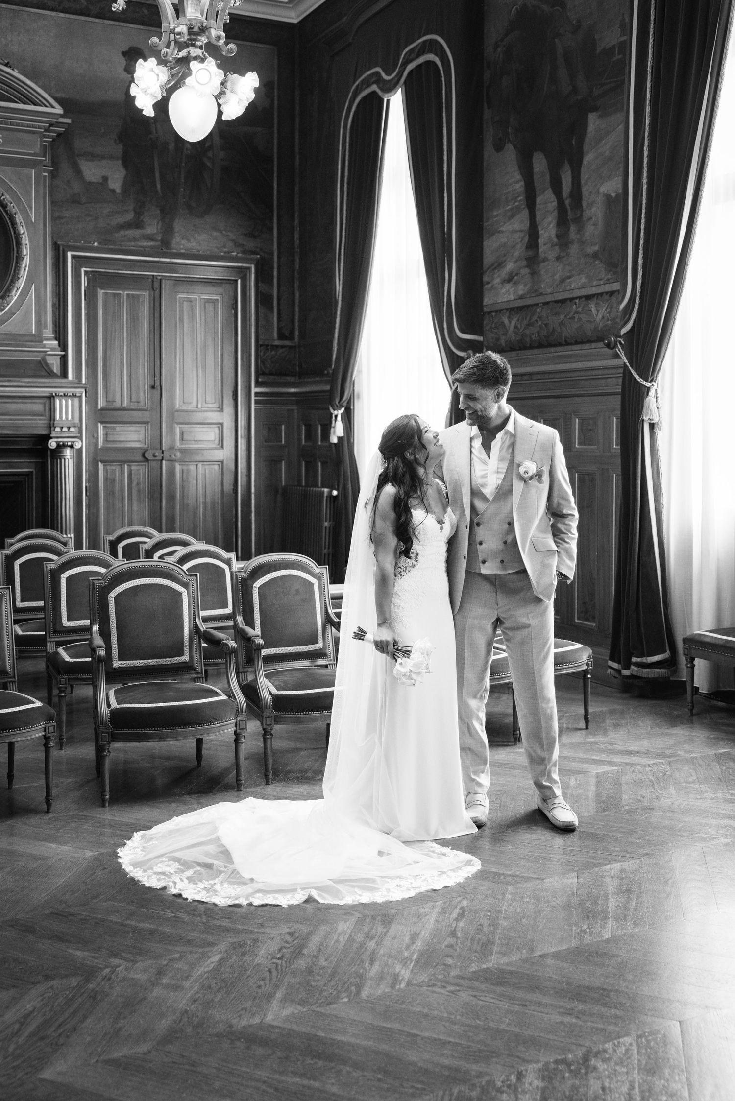 Photo en noir et blanc d'une mariée et d'un mari partageant un moment calme dans une salle de cérémonie élégante avec une longue traîne