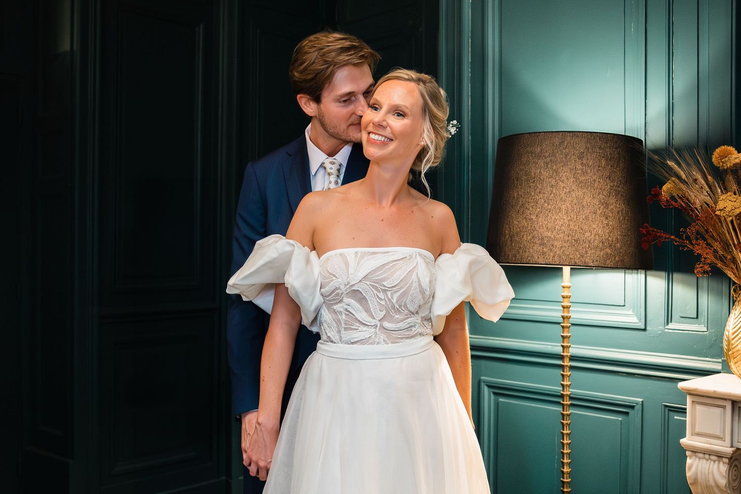 Groom in a navy suit embraces a smiling bride in an off-shoulder wedding gown against a deep green wall.