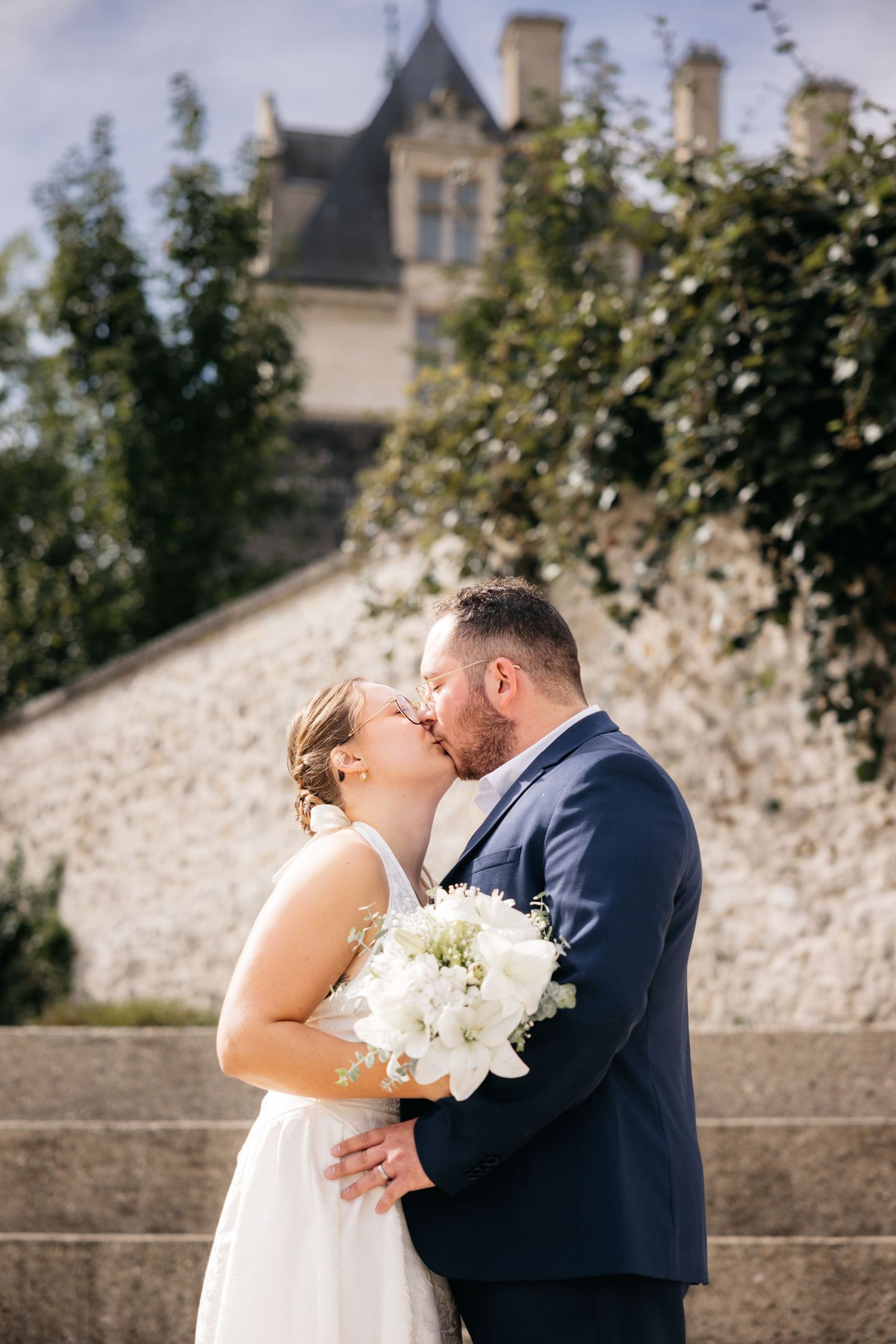 Bride in a white dress and groom in a navy suit kiss on stone steps with a bouquet, an ivy wall and a castle behind.