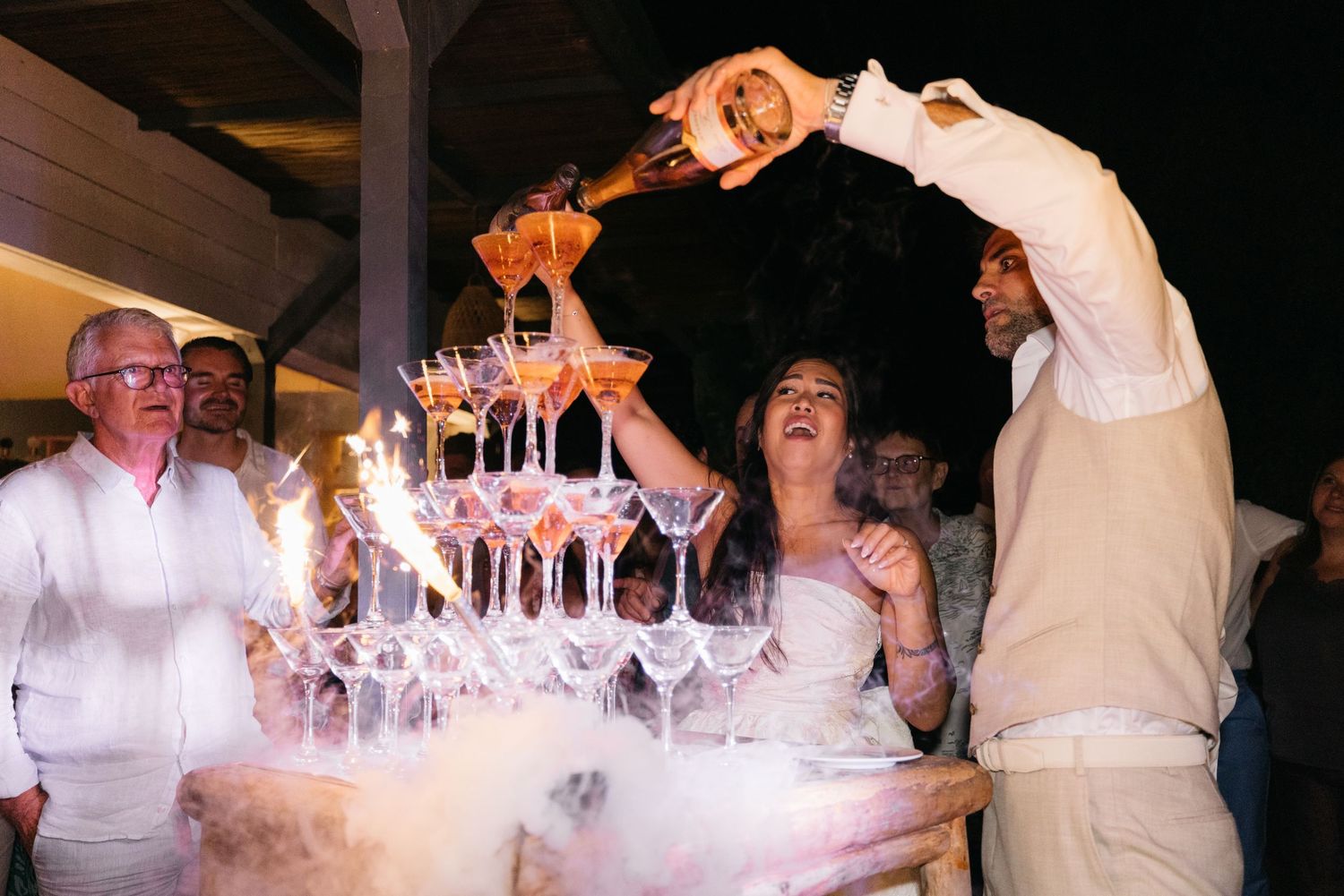 Groom pours champagne over a tall champagne tower as the laughing bride and guests watch at an evening wedding celebration.