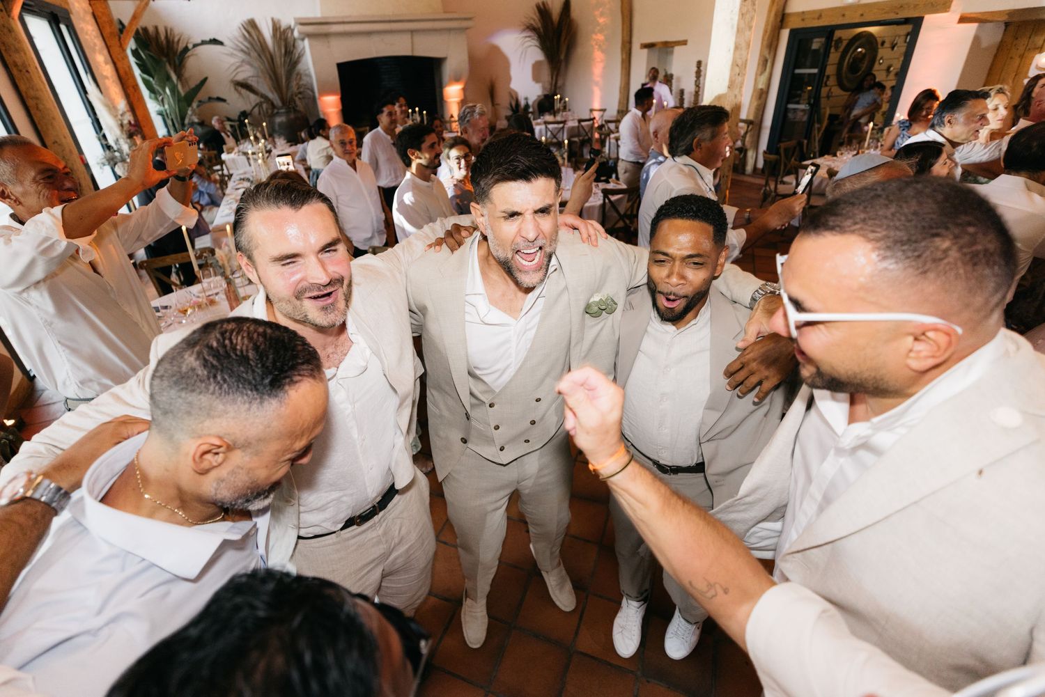 Groomsmen in cream suits laugh and celebrate together on the dance floor during a lively wedding reception.