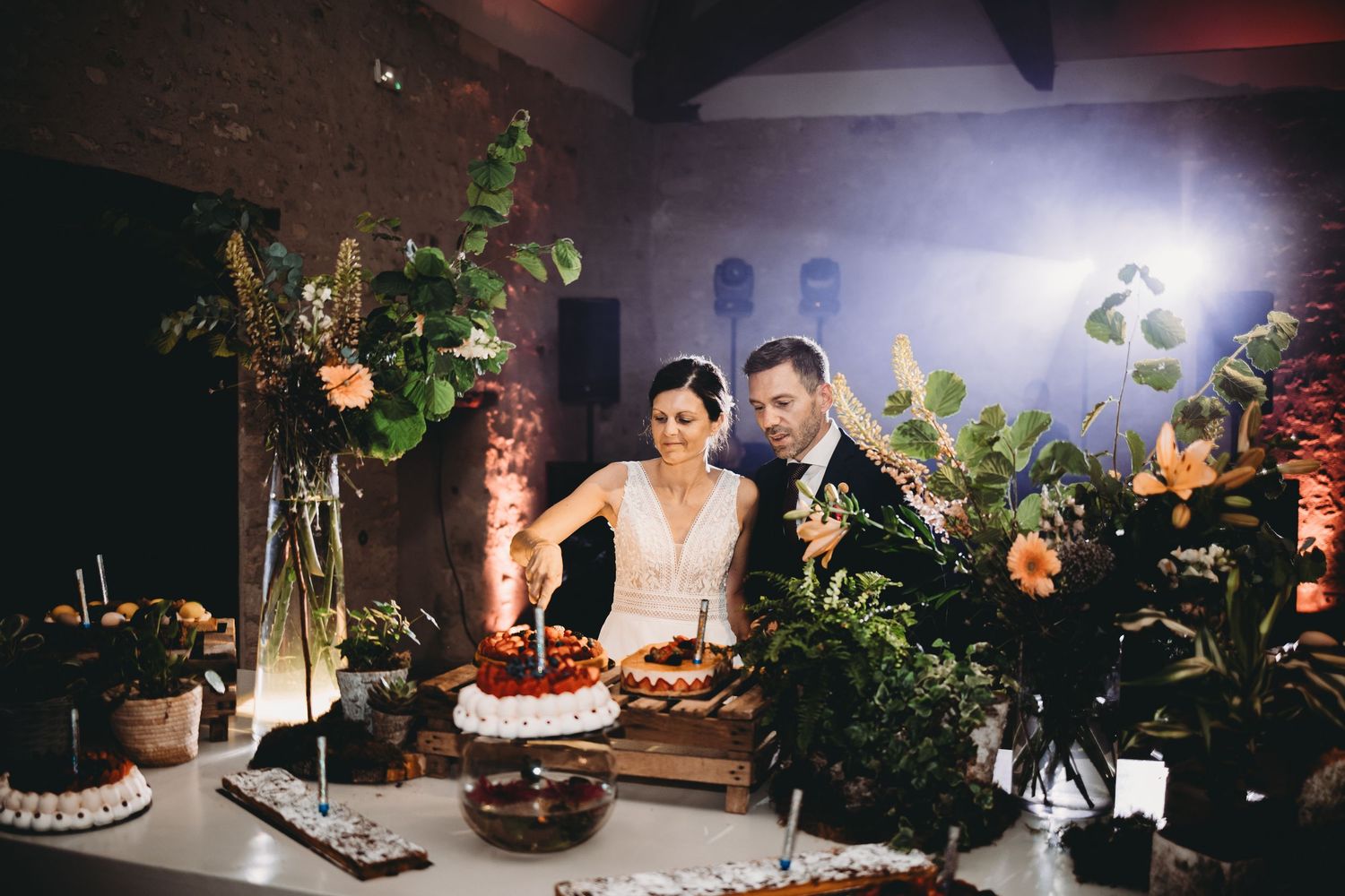 Bride and groom cut a tiered wedding cake together at a rustic reception, surrounded by lush floral arrangements and warm ambient lighting.
