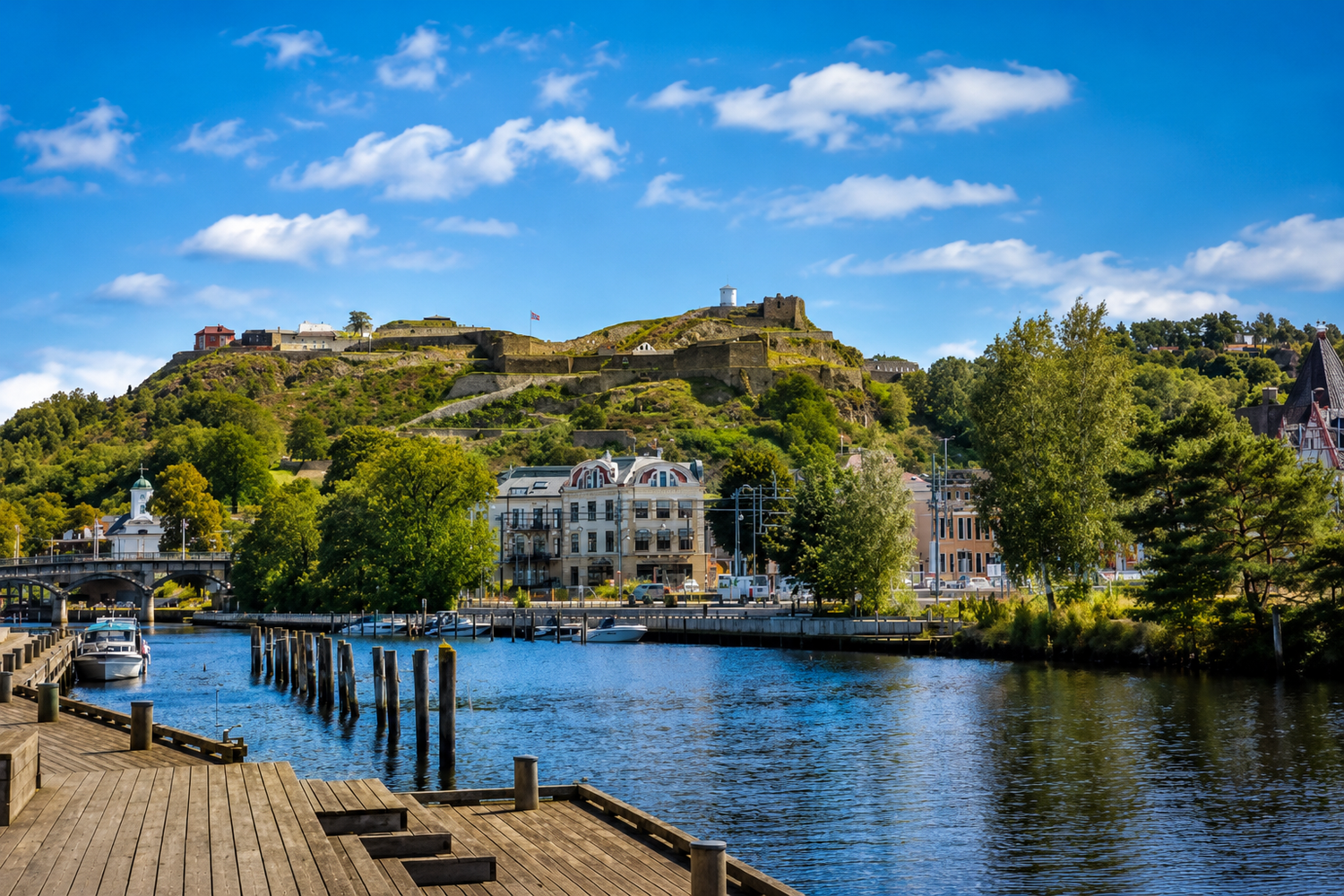 Sunny harbor with a wooden dock, calm blue water, and a hilltop fortress rising above green trees and historic buildings.