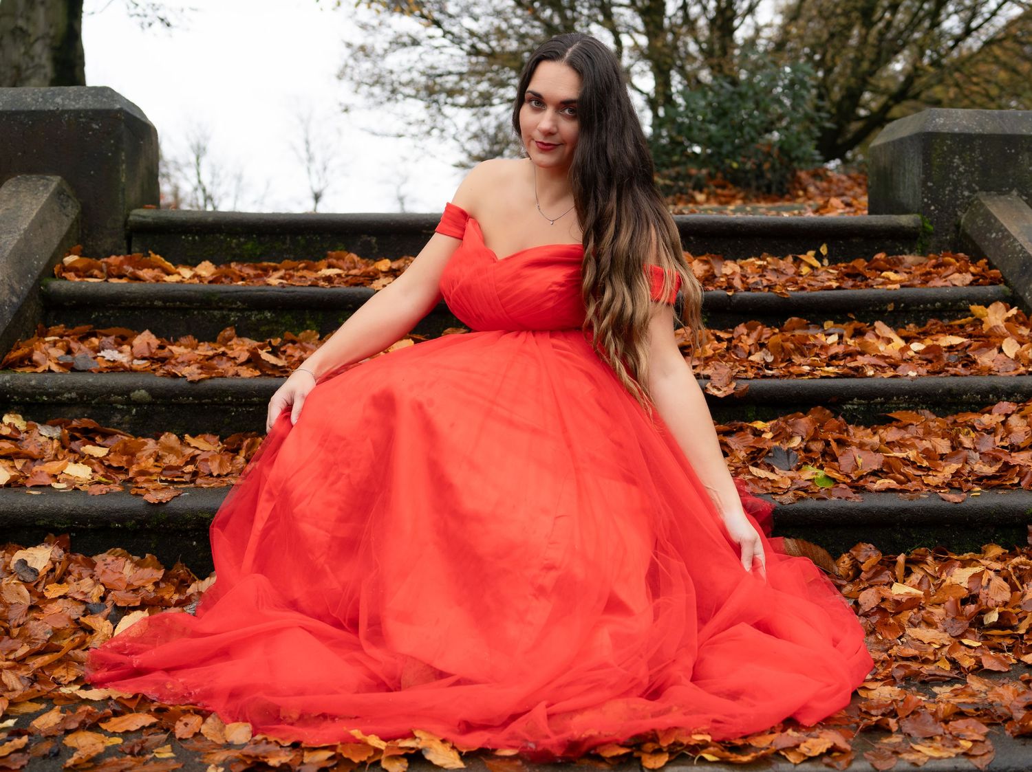Figure in a flowing red dress posed among autumn leaves on the ground.