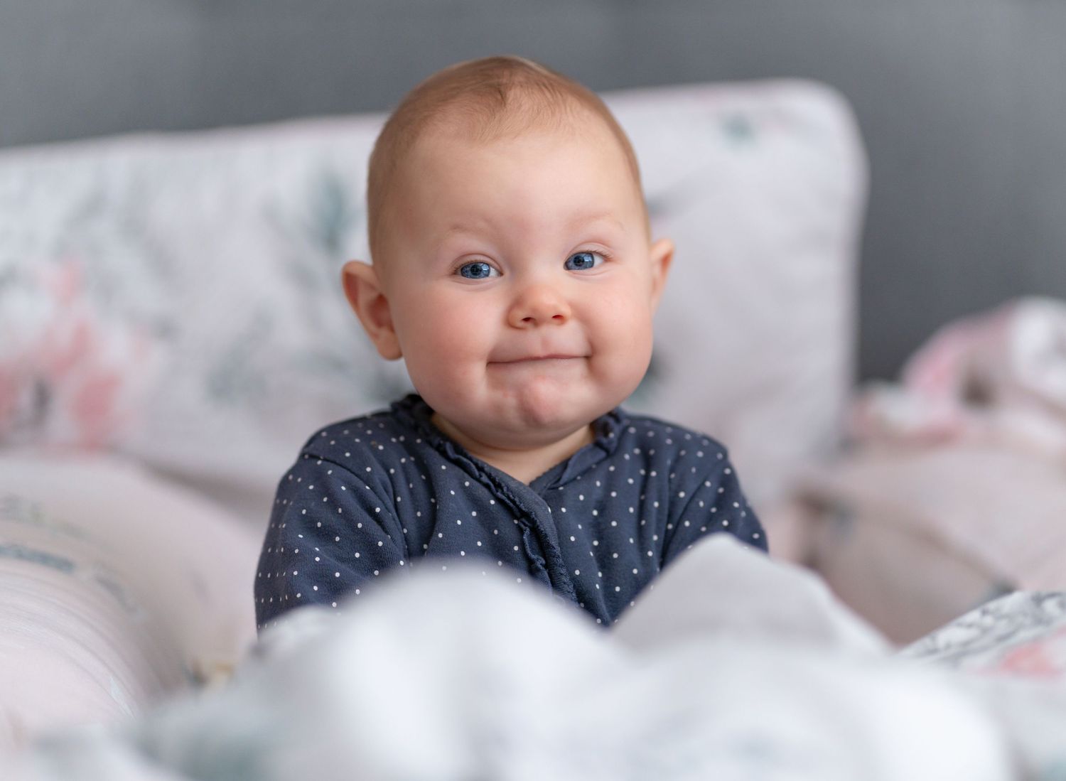 A baby in a navy polka dot outfit smiles while lying on white bedding.