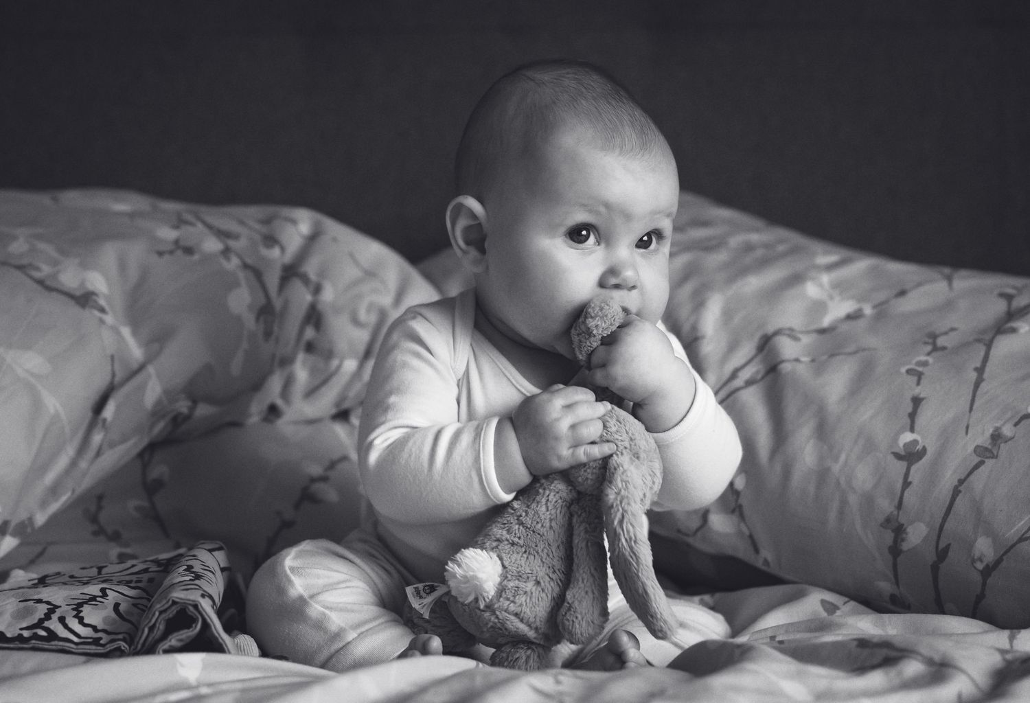 Black and white photo of baby in white onesie sitting on bed with toy.