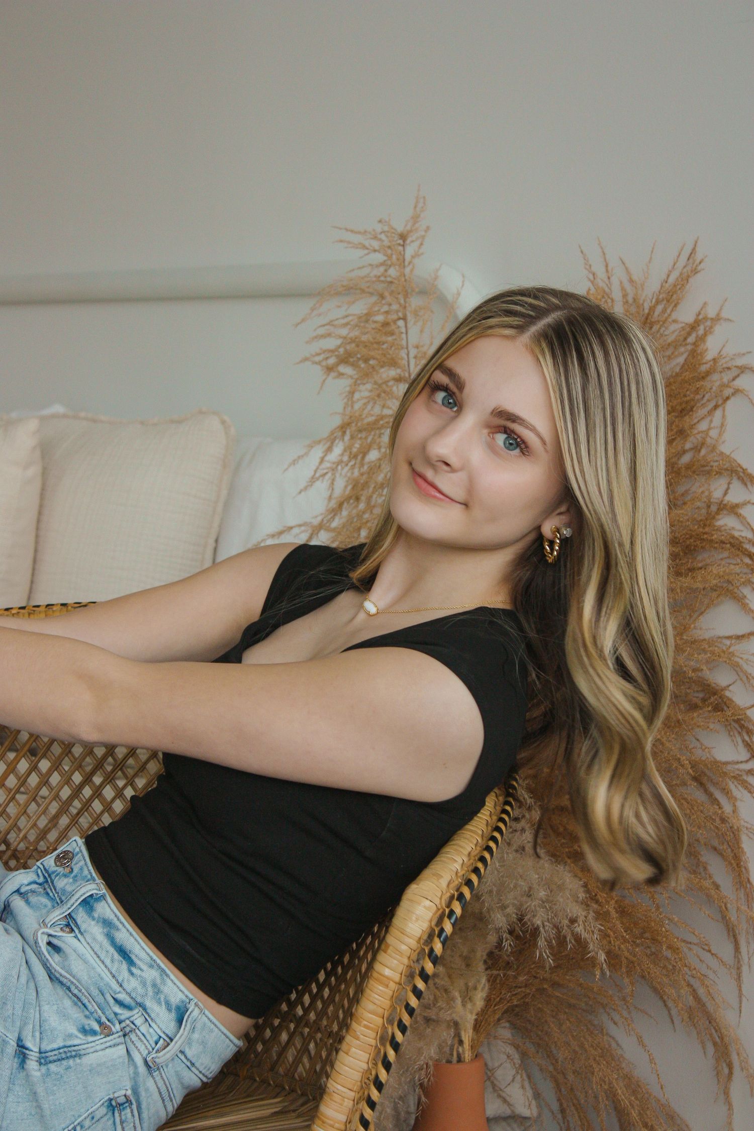Young blonde woman in black top and denim shorts relaxing in wicker chair with pampas grass decor.