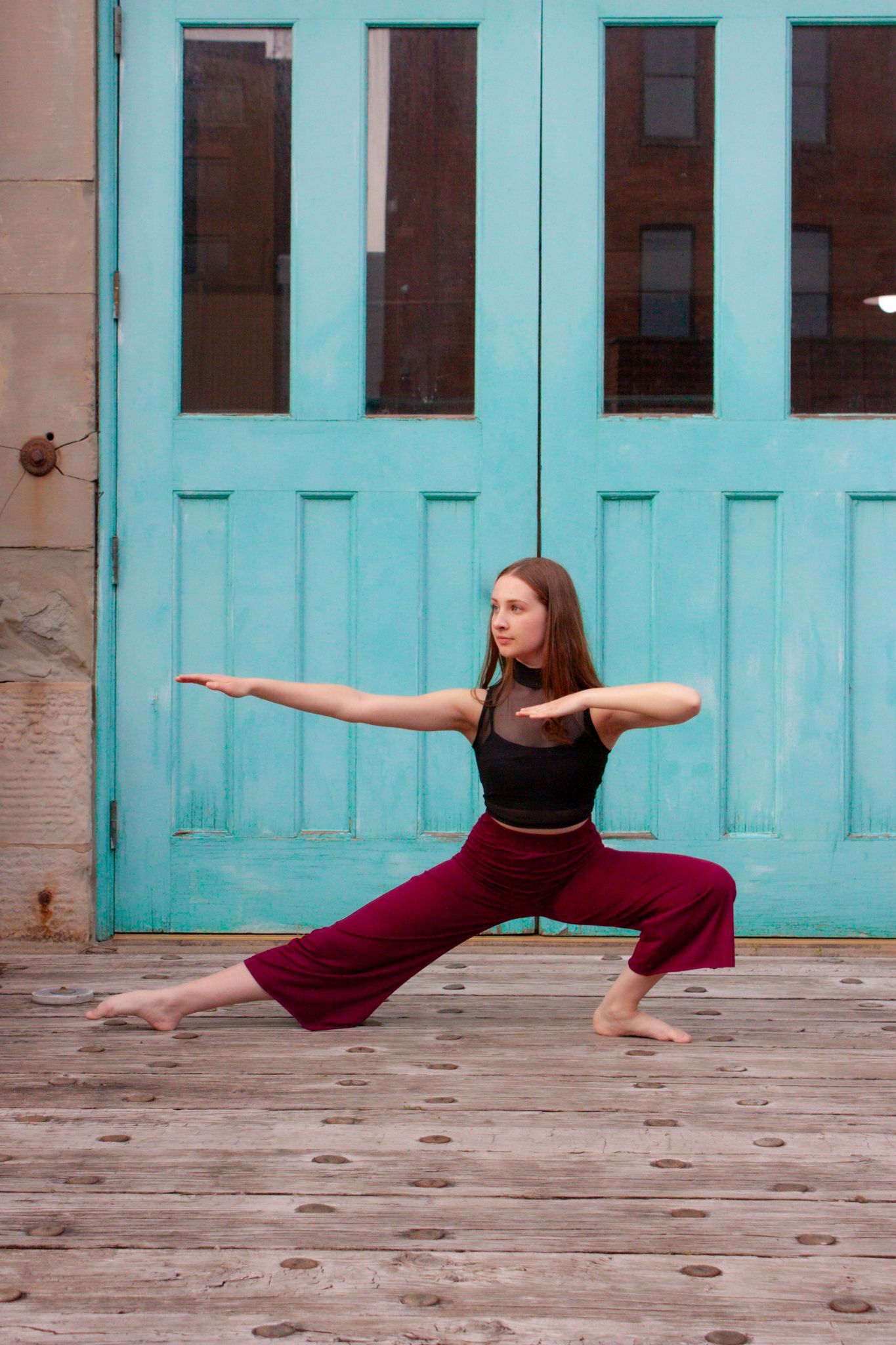 Female dancer in warrior pose wearing burgundy wide-leg pants and black top on wooden deck before turquoise doors.