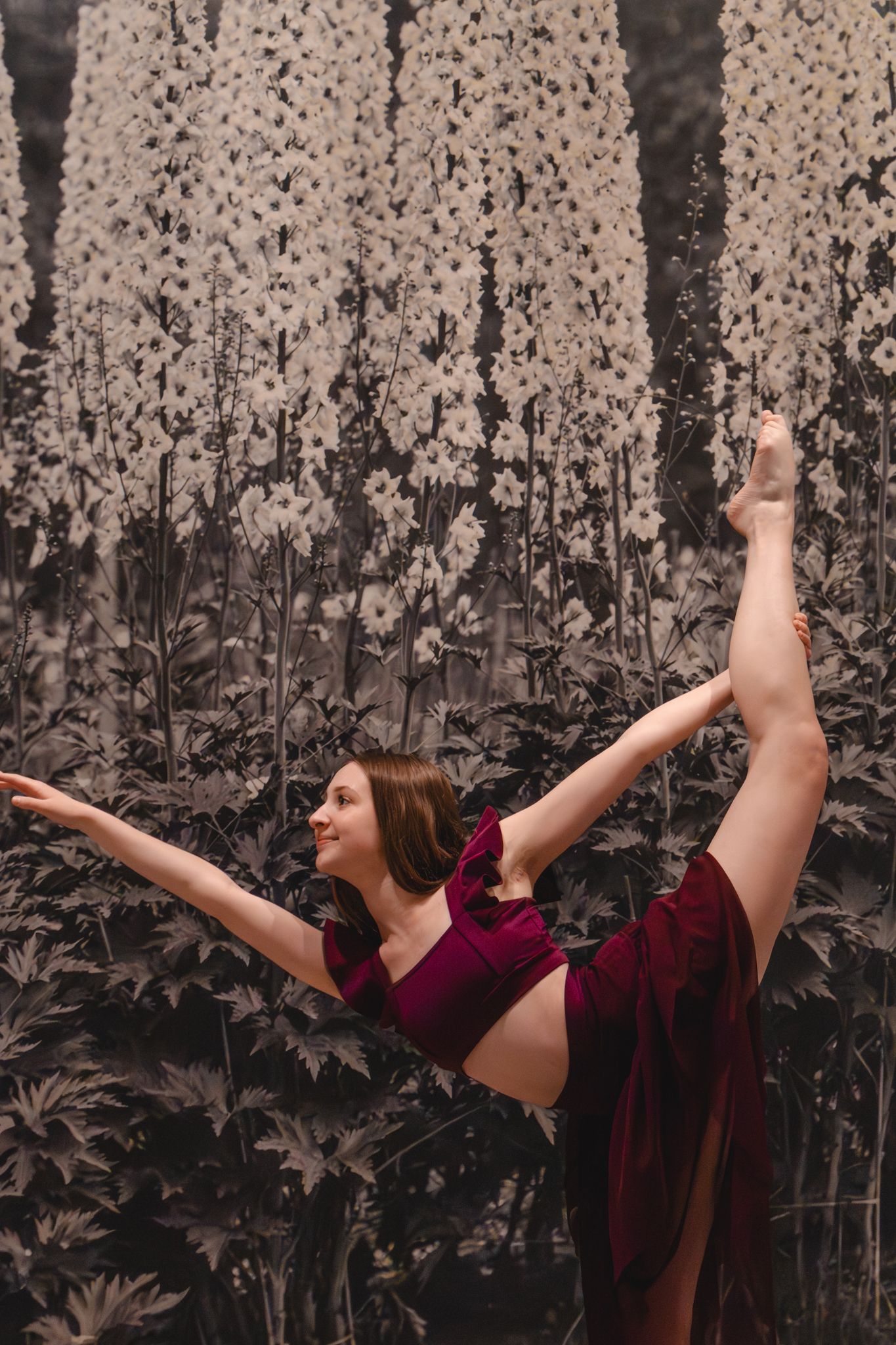 A flexible dancer in a burgundy outfit performs a standing split pose against a monochrome floral background.