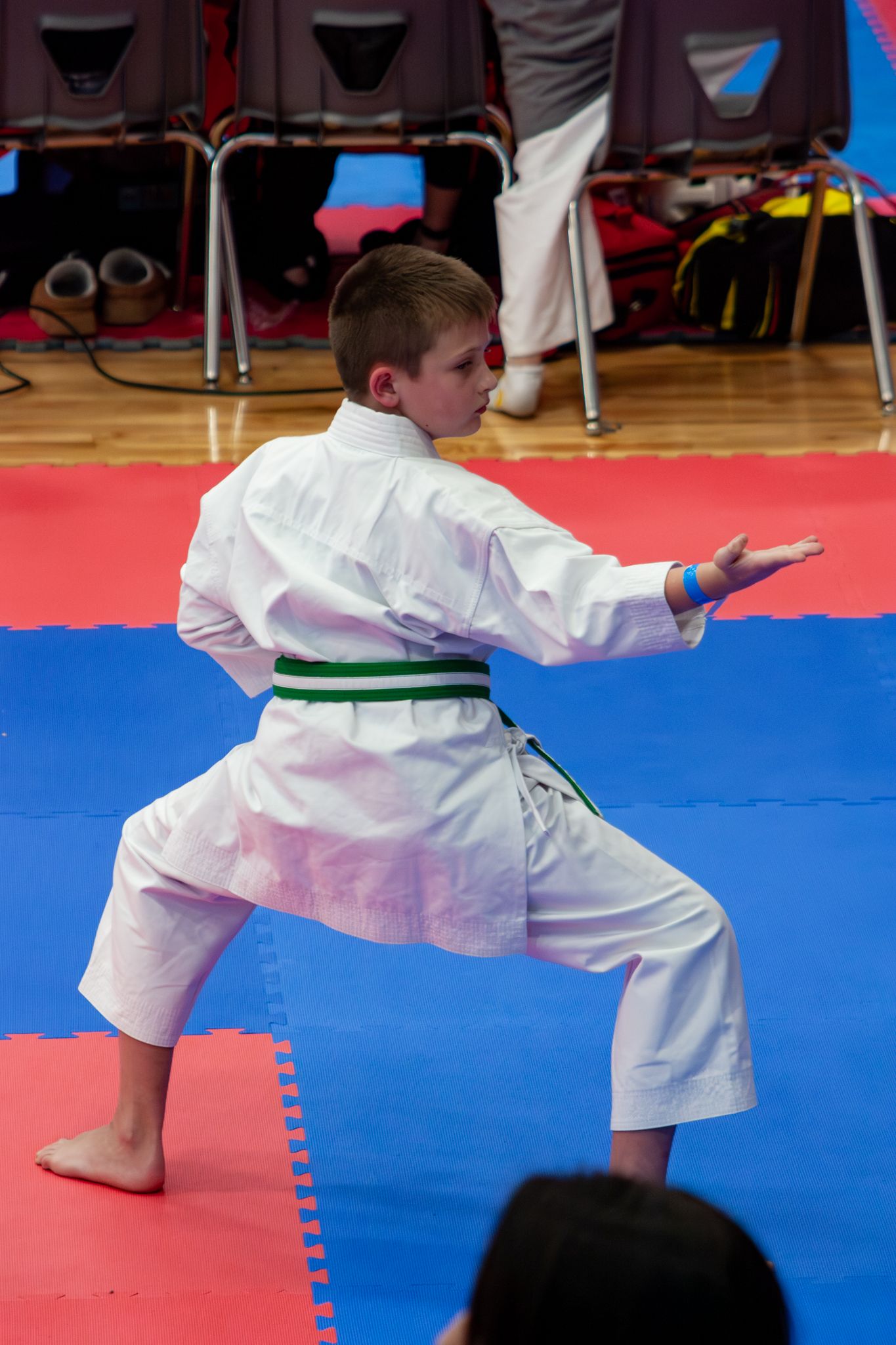 Young karate student in white gi with green belt executes a precise kata technique on red and blue competition mats.